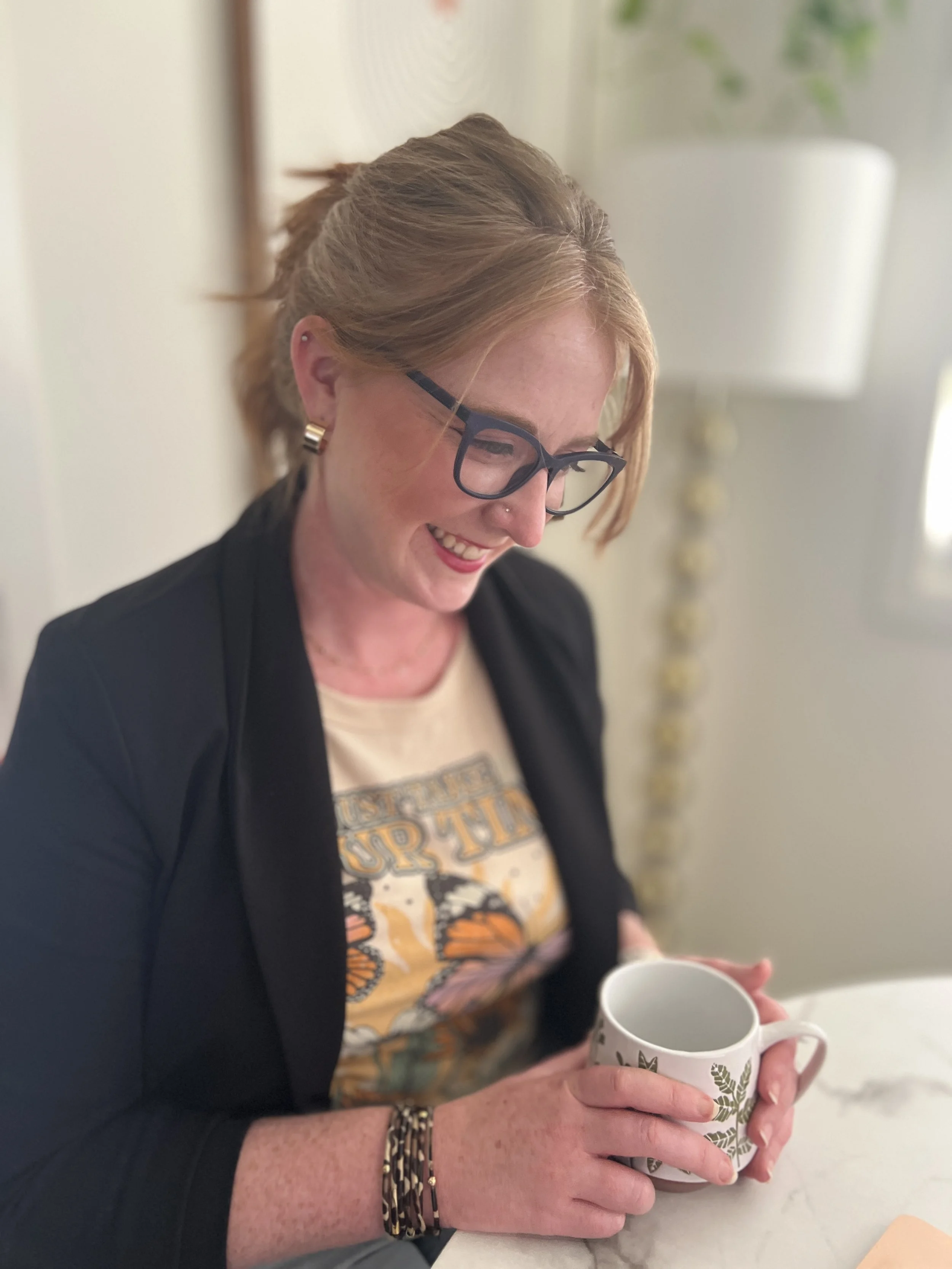 A woman with light brown hair, wearing glasses and a black blazer, is holding a white mug with green leaf patterns. She is smiling and looking down. In the background, there is a white lamp and a plant.