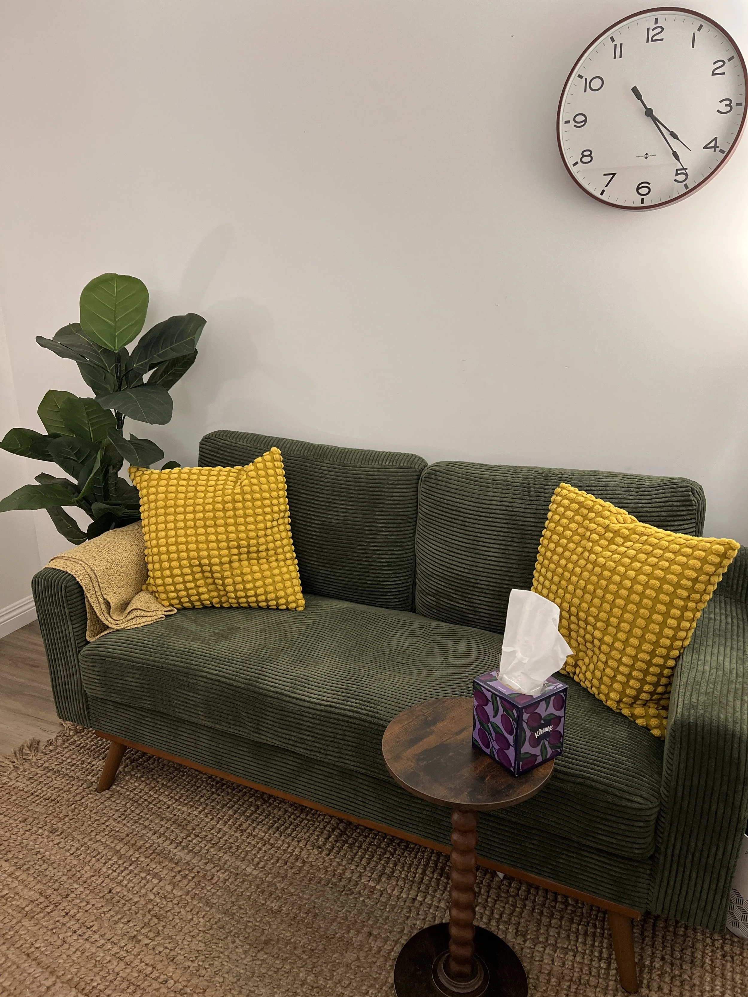 A living room waiting area with a green striped sofa, yellow textured pillows, a wooden coffee table with a tissue box, a potted plant, a wall clock, and a beige rug.