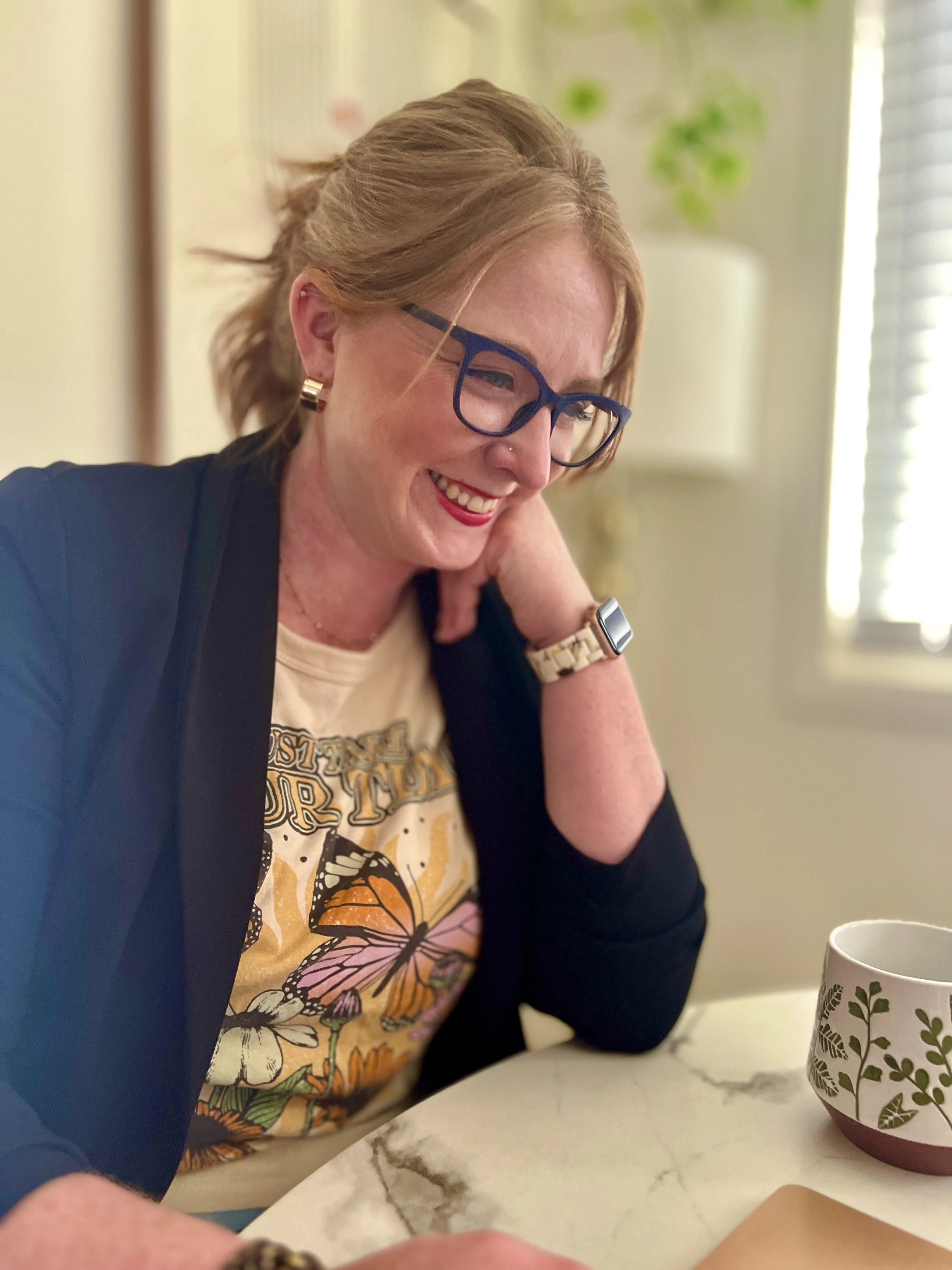 A woman with glasses and red hair, smiling, wearing a butterfly graphic t-shirt, and sitting at a table with a plant-patterned mug in front of her.