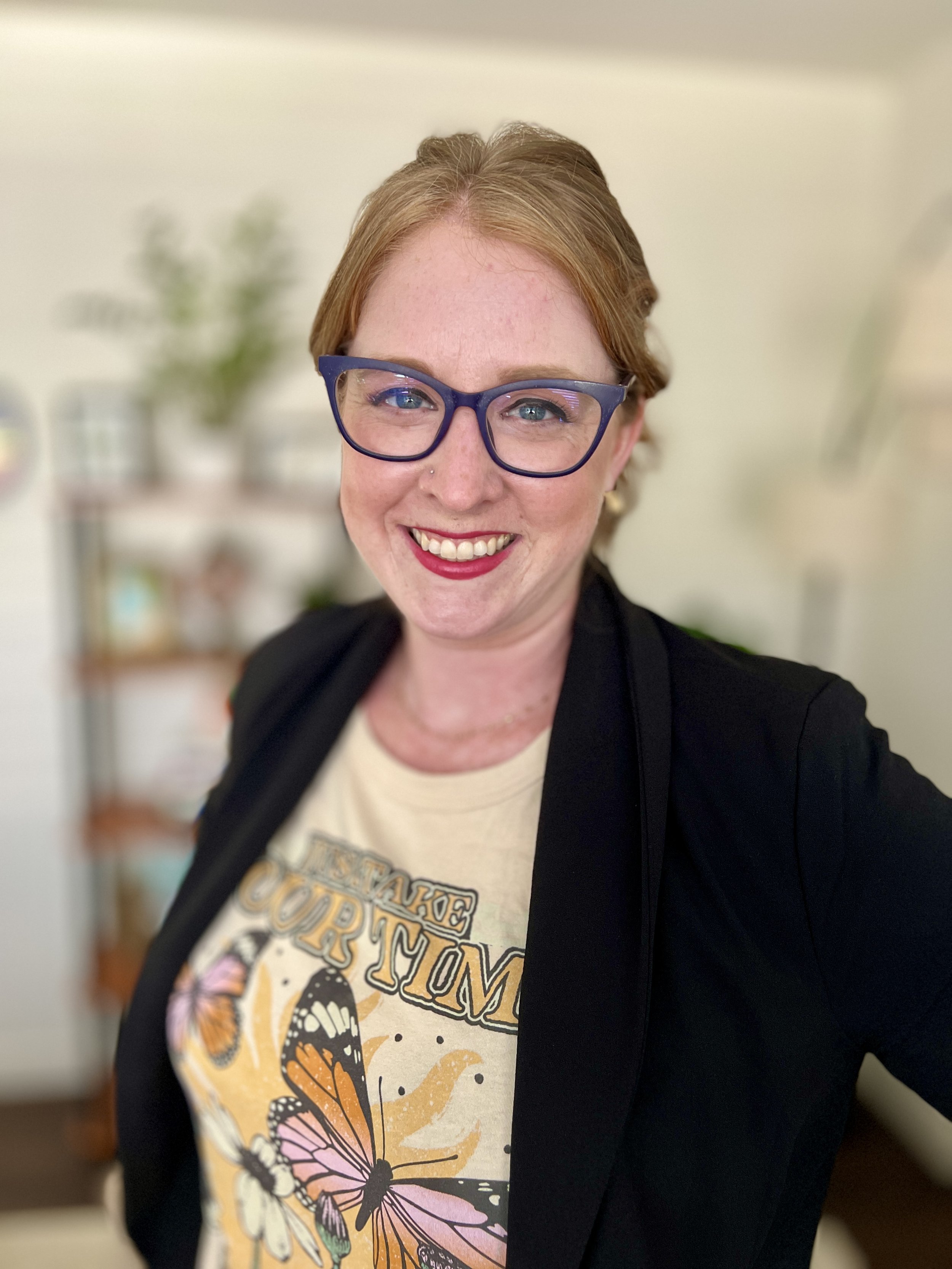 Close-up of a woman with red hair, glasses, and a nose piercing smiling in an indoor setting.