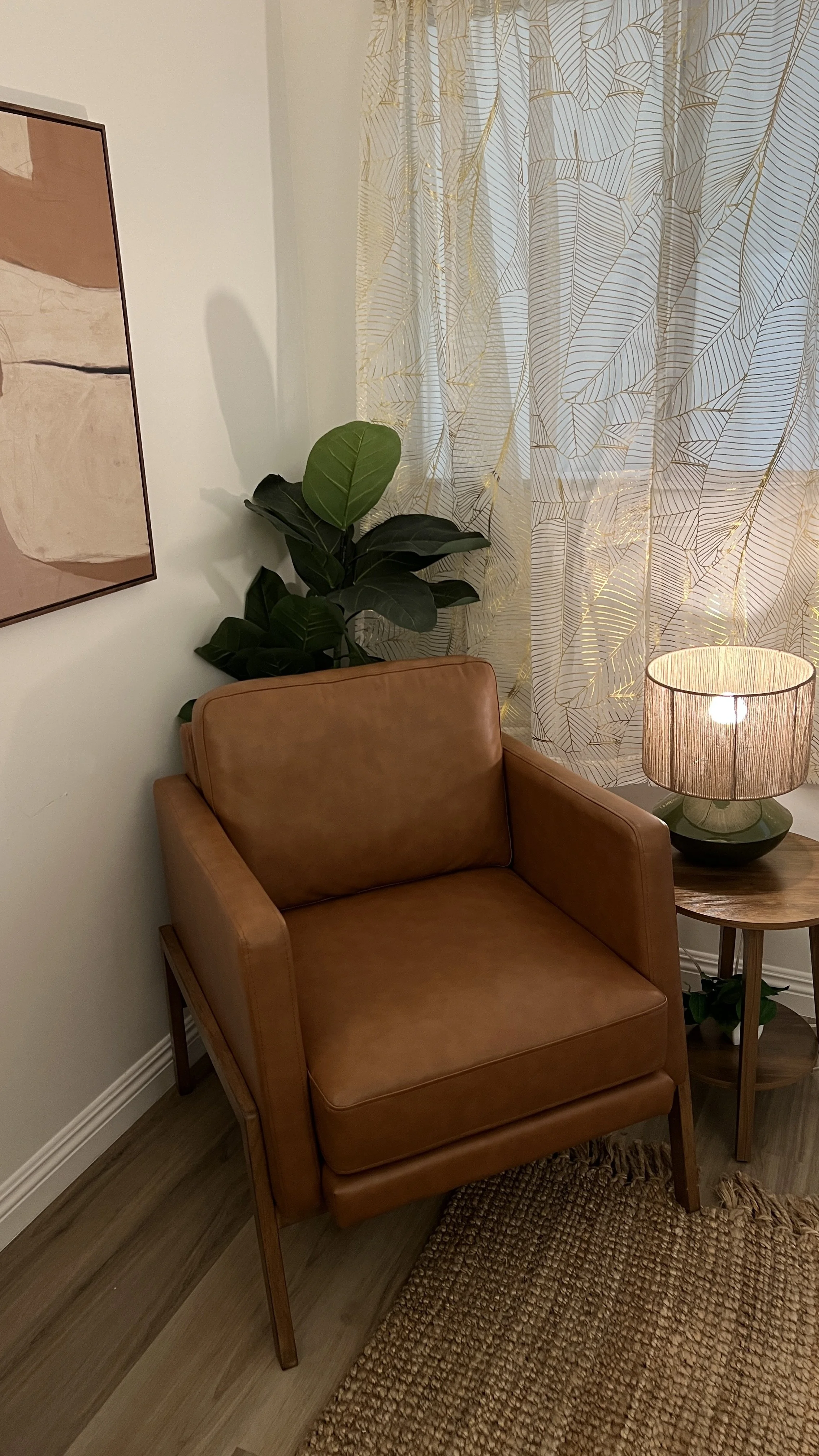 A cozy living room corner with a tan leather armchair, a small wooden side table with a table lamp, a green houseplant with large leaves, a dresser with art on the wall, and sheer curtains with gold patterns.