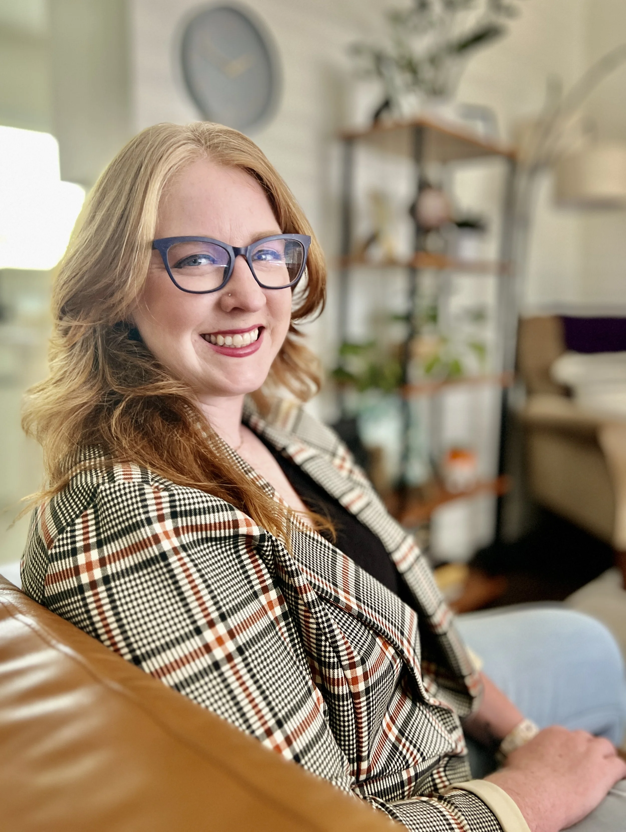A woman with red hair and glasses smiling, sitting indoors with a bookshelf and a clock in the background.