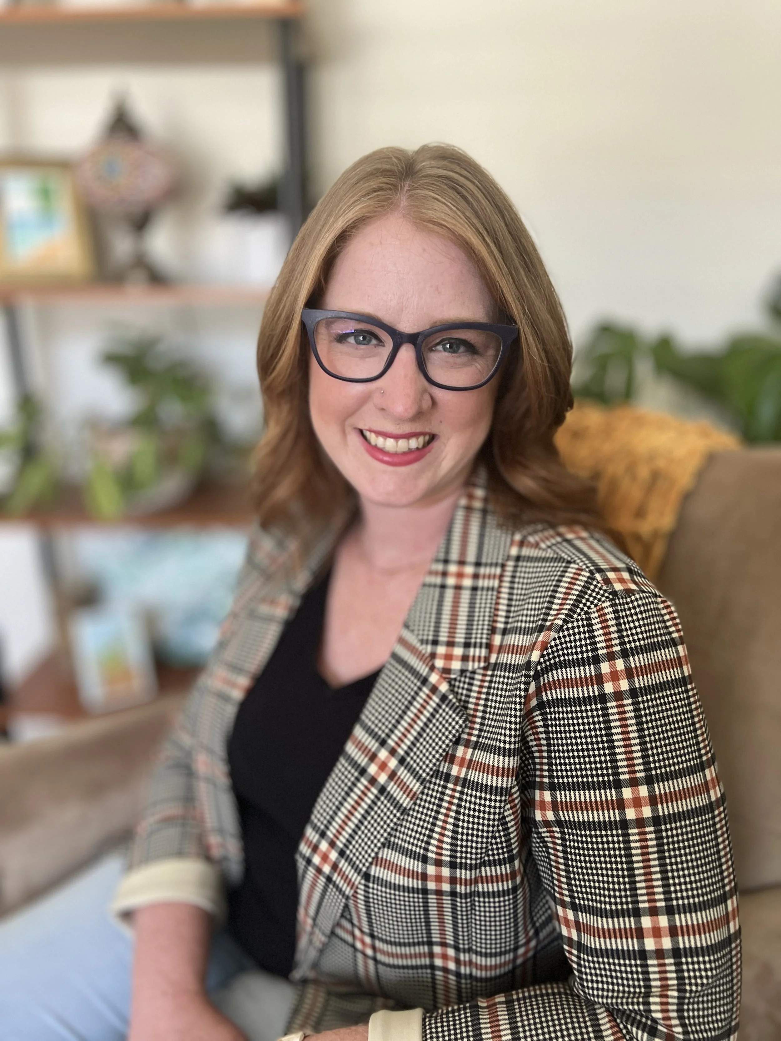 A woman with glasses and a plaid blazer smiling with a blurred indoor background.