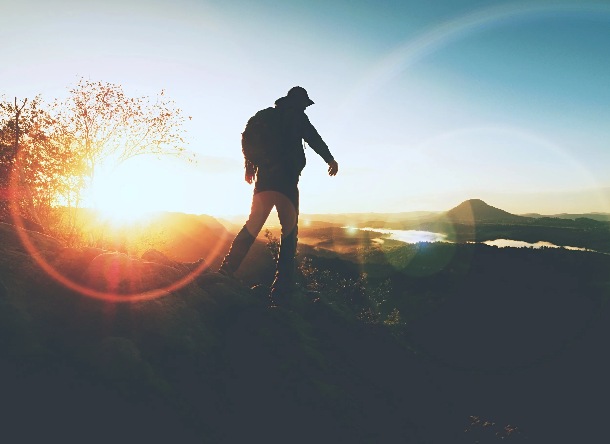 Silhouette of a person hiking on a mountain at sunset, with trees, a lake, and distant hills in the background.