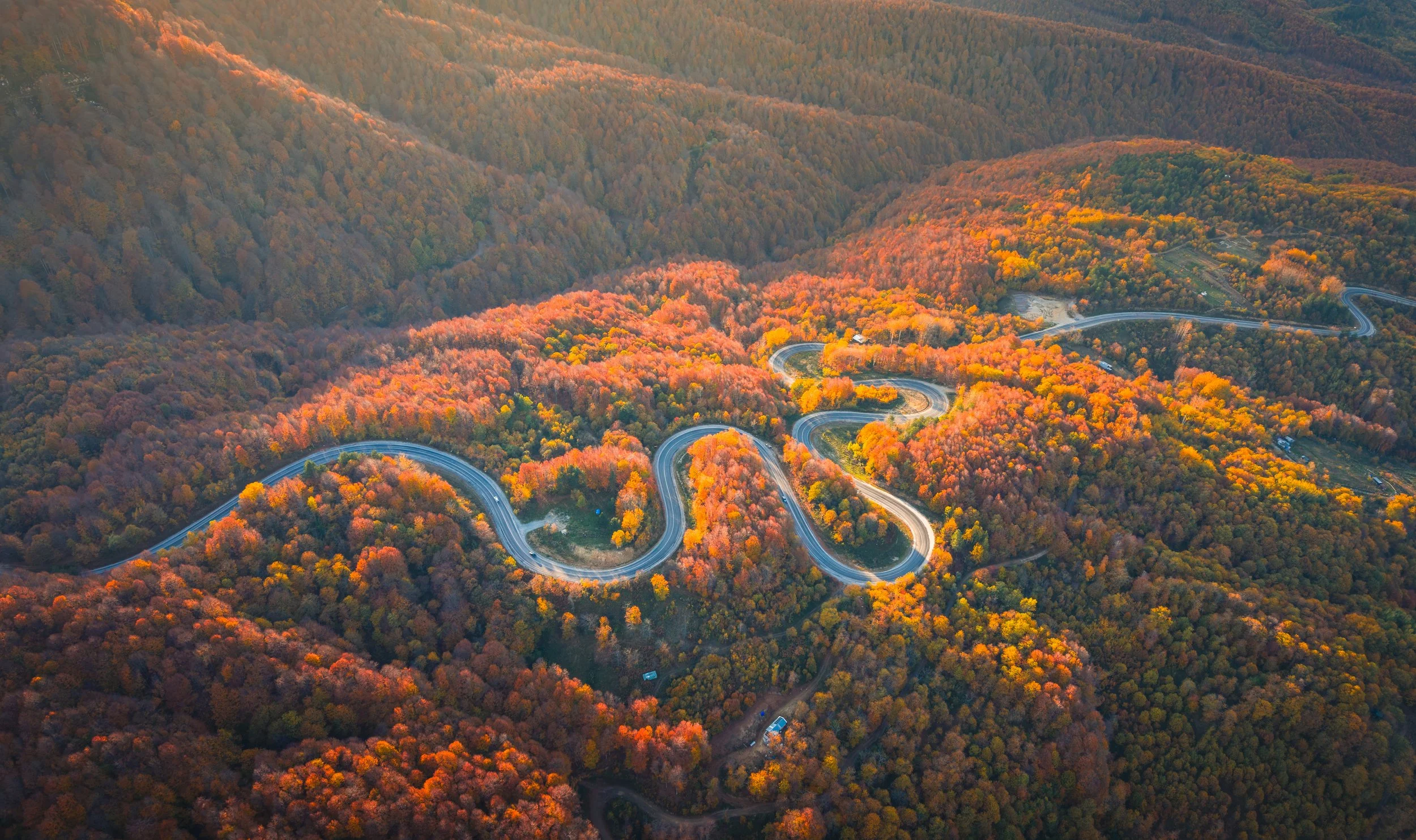 An aerial view of a winding mountain road through a forest with autumn foliage in shades of orange, yellow, and red.