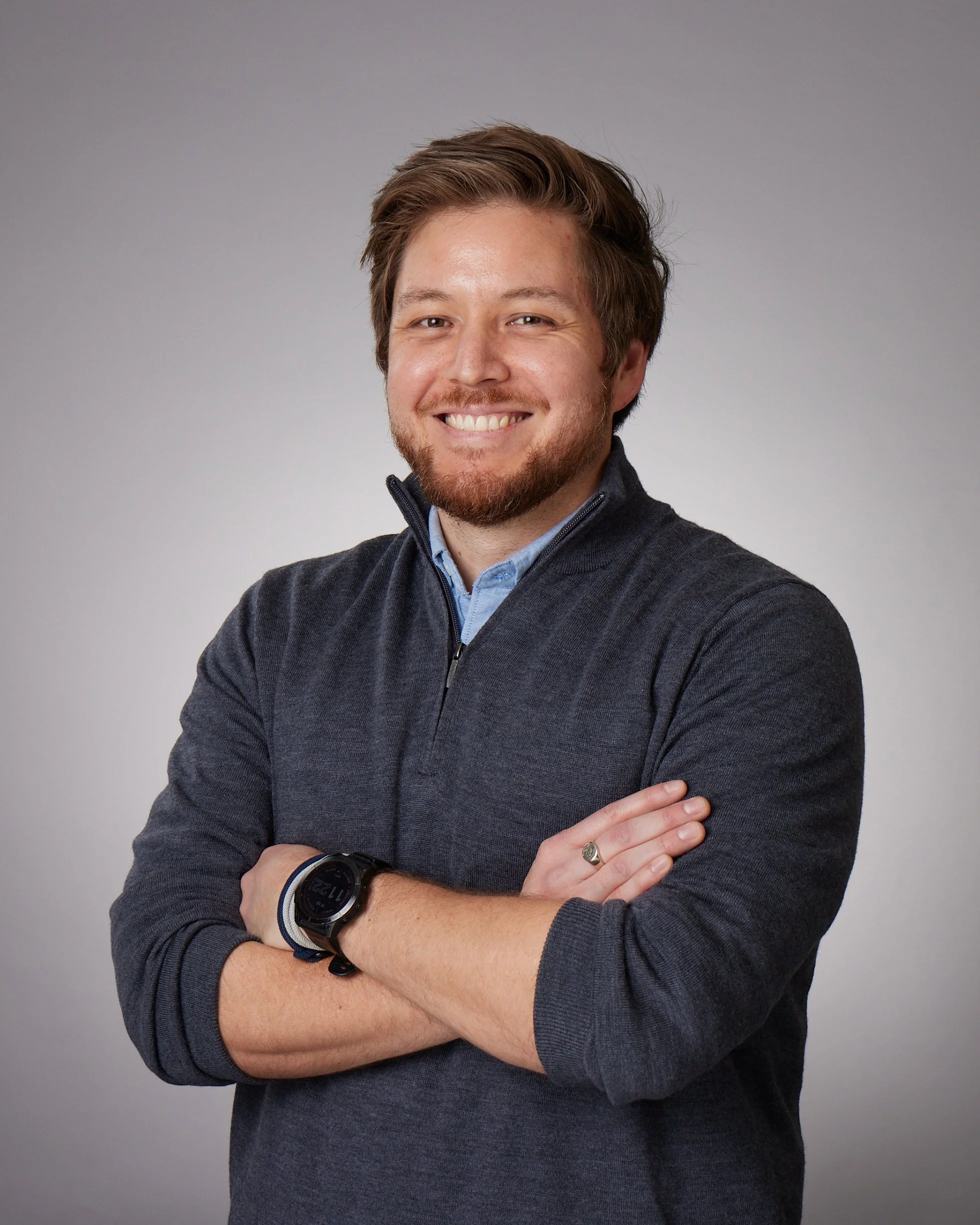 A man with brown hair and a beard, smiling, wearing a dark gray fleece jacket over a blue shirt, standing with his arms crossed against a gray background.