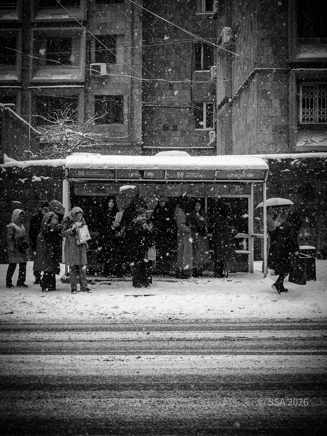 People waiting at a bus stop in snowy weather with snow falling and buildings in the background.