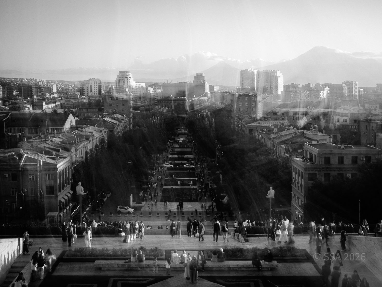View of a busy urban area with a large staircase leading down to a street filled with many people walking. The scene includes various buildings, trees, and mountains in the distance, with a hazy sky.