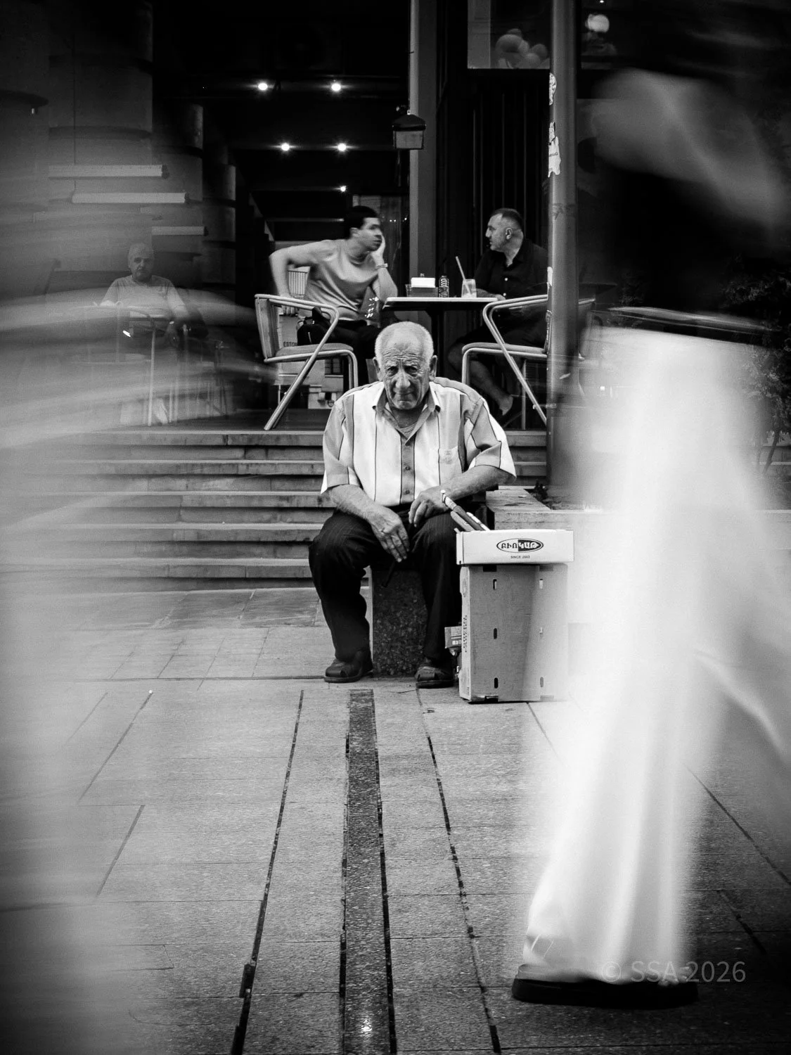 An elderly man with a cane sitting on a small box, looking at the camera, outside a café or restaurant. In the background, two young men are seated at a table, engaged in conversation. The scene is black and white, with motion blur of pedestrians passing by.