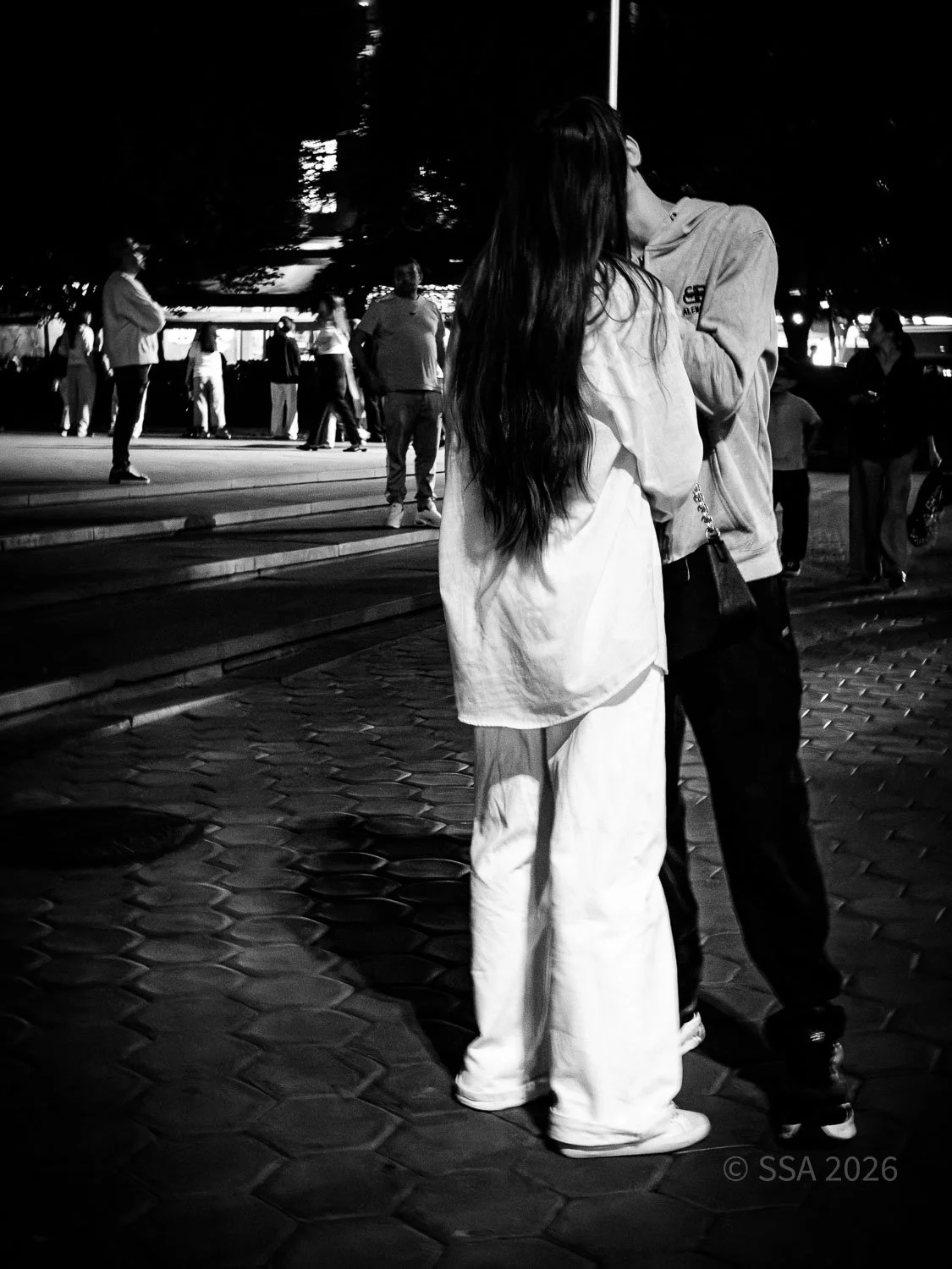 Black and white photo of a couple kissing, standing on a brick sidewalk at night. Several people are in the background, some standing and some walking, with trees and lights visible in the distance.