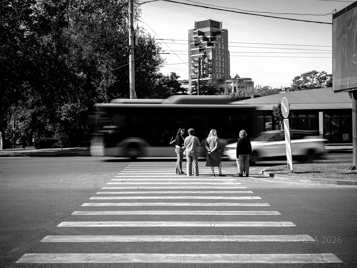 A black-and-white photo of a city street showing four women standing on a crosswalk waiting to cross, with a blurred bus and car passing by. There are trees and tall buildings in the background.