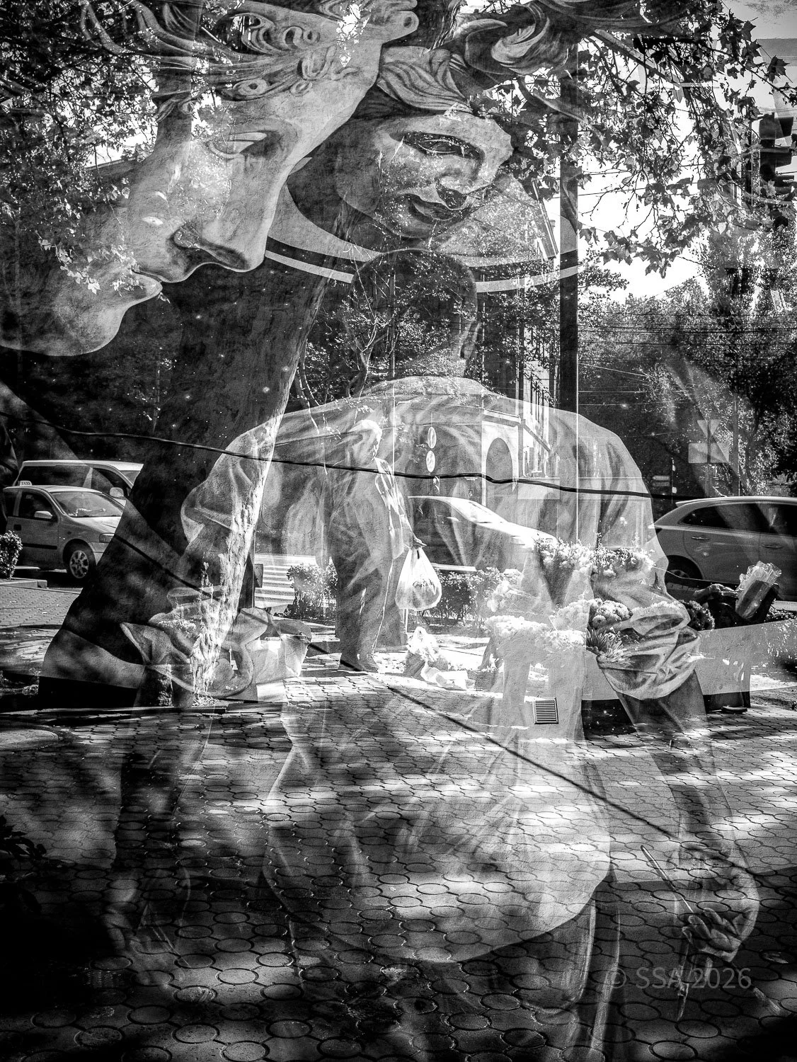 Black and white photo of a reflection showing a statue of two women, one with flowers in her hair, and people on the street with parked cars and buildings in the background.