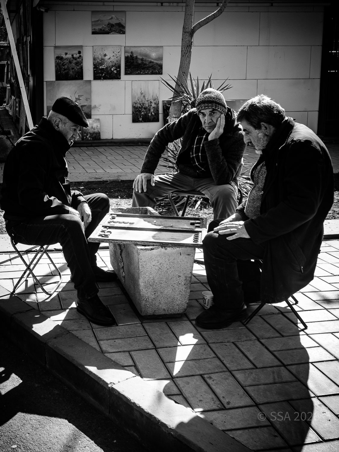 Three men sitting around a table playing checkers outdoors on a sunny day, with art photos on the wall behind them.