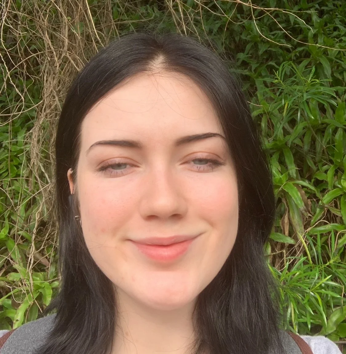 A young woman with dark hair and light skin, smiling with slightly squinted eyes, standing outdoors in front of green leafy plants and brown vines.