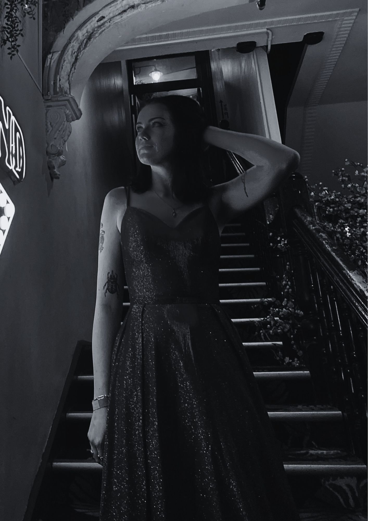 Black and white photo of a woman with dark hair and tattoos on her arm, wearing a sparkly dress, posing with her left hand behind her head inside a restaurant or bar with ornate decor and a window in the background.