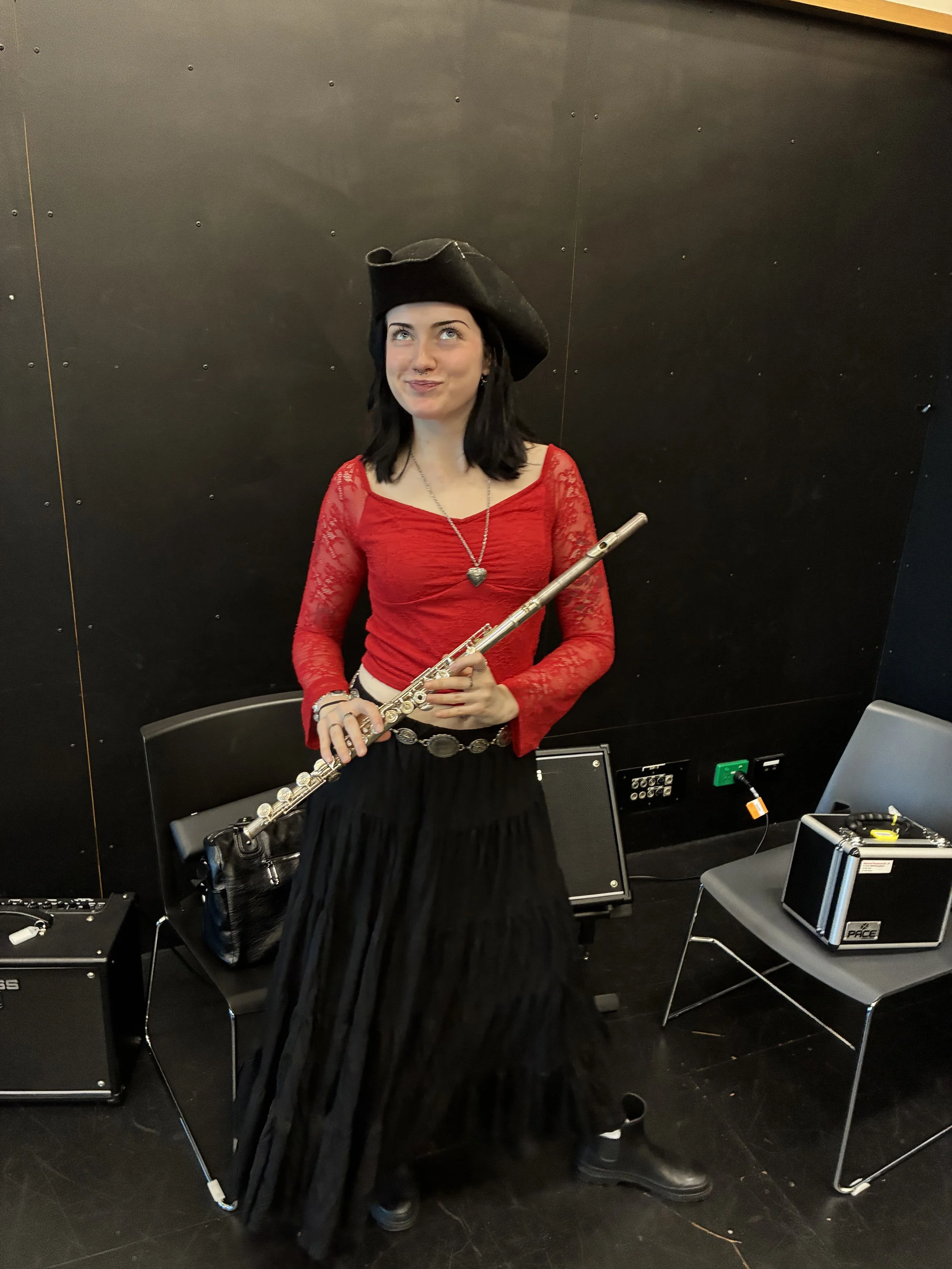A woman dressed in a red lace top, black skirt, and pirate hat holding a flute in a room with black walls and musical equipment.