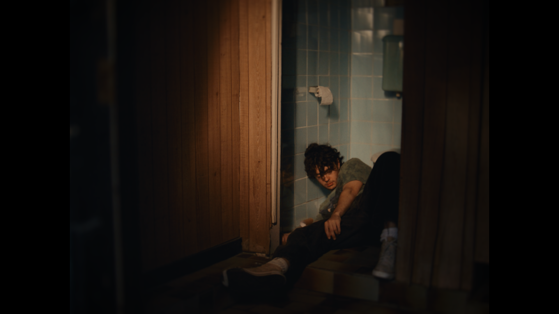 A person with curly hair and glasses lying on the floor of a bathroom in a corner, wearing a t-shirt and pants, with a distressed expression, seen through a slightly opened door.