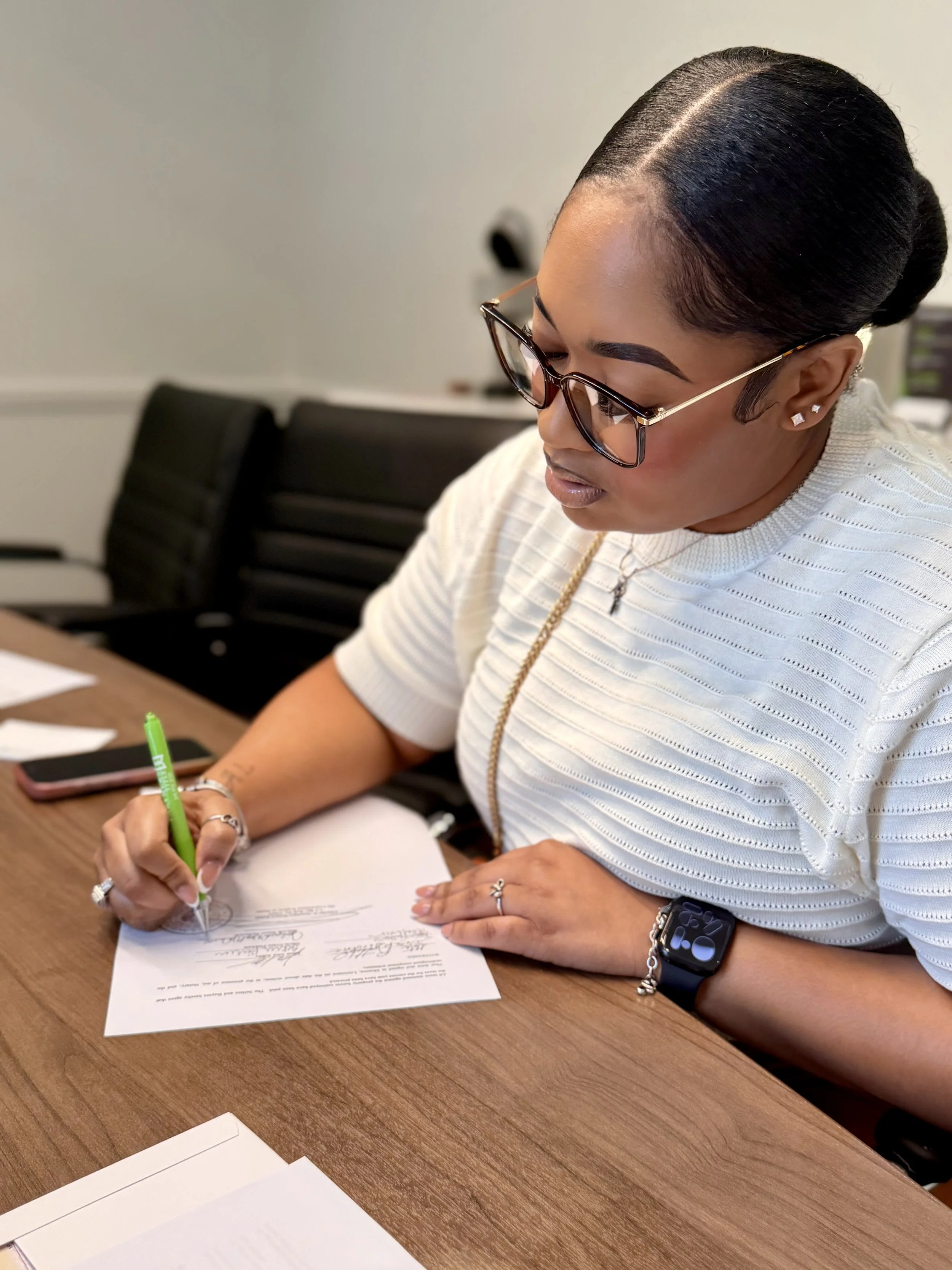 A woman with dark hair, glasses, and jewelry is sitting at a wooden table, signing a document with a green pen. There are loose sheets of paper on the table.