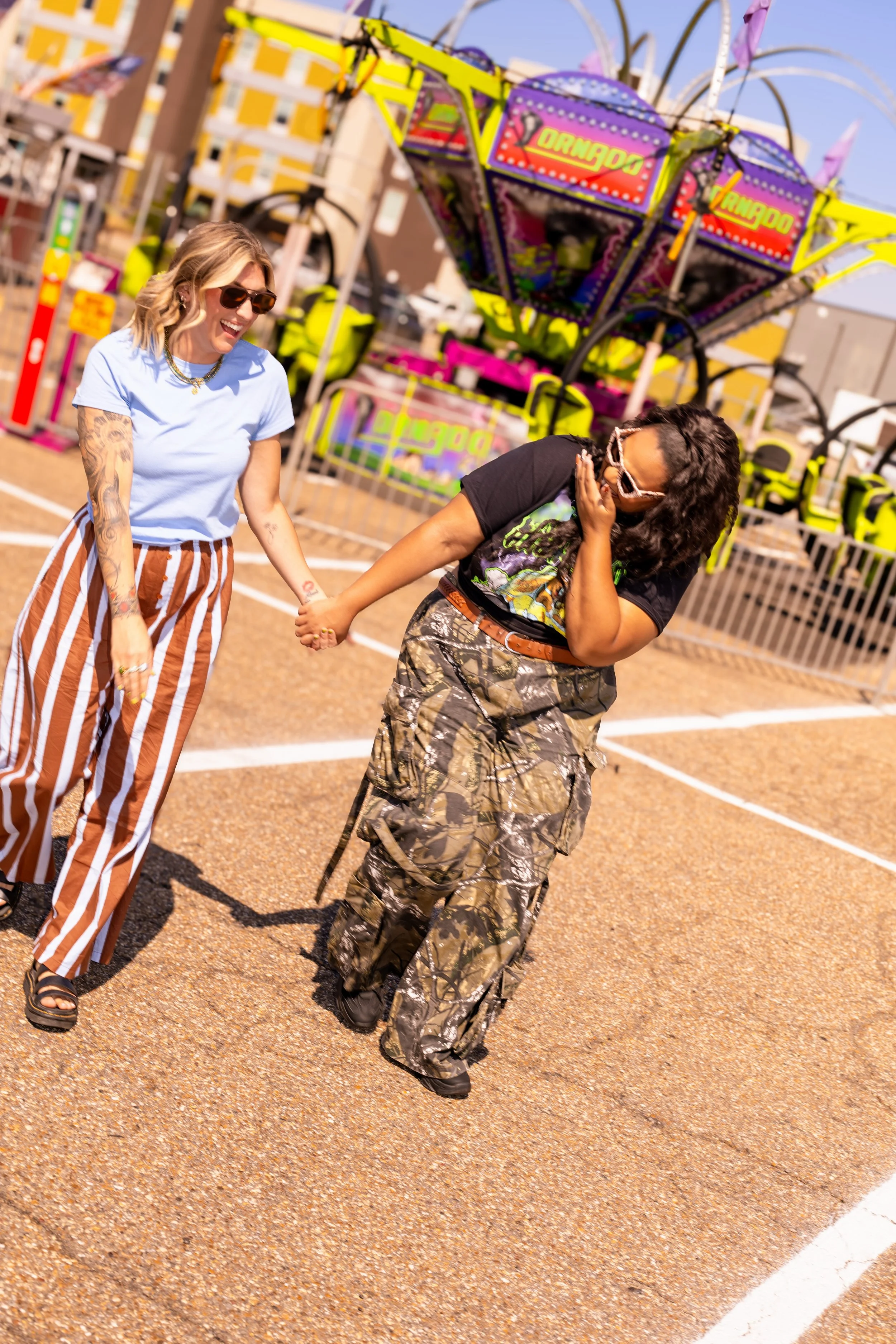 Two women holding hands and laughing at an outdoor amusement park with a colorful ride in the background.