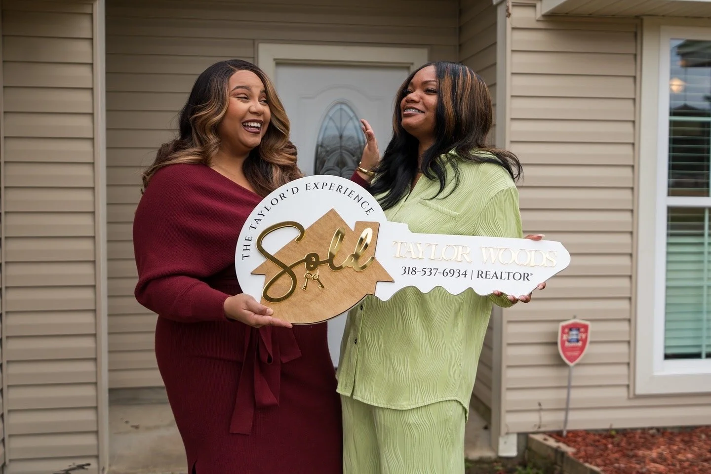 Two women smiling and holding a sign that reads "The Taylor Experience, Sold, Taylor Woods." They stand in front of a house, celebrating a real estate transaction.