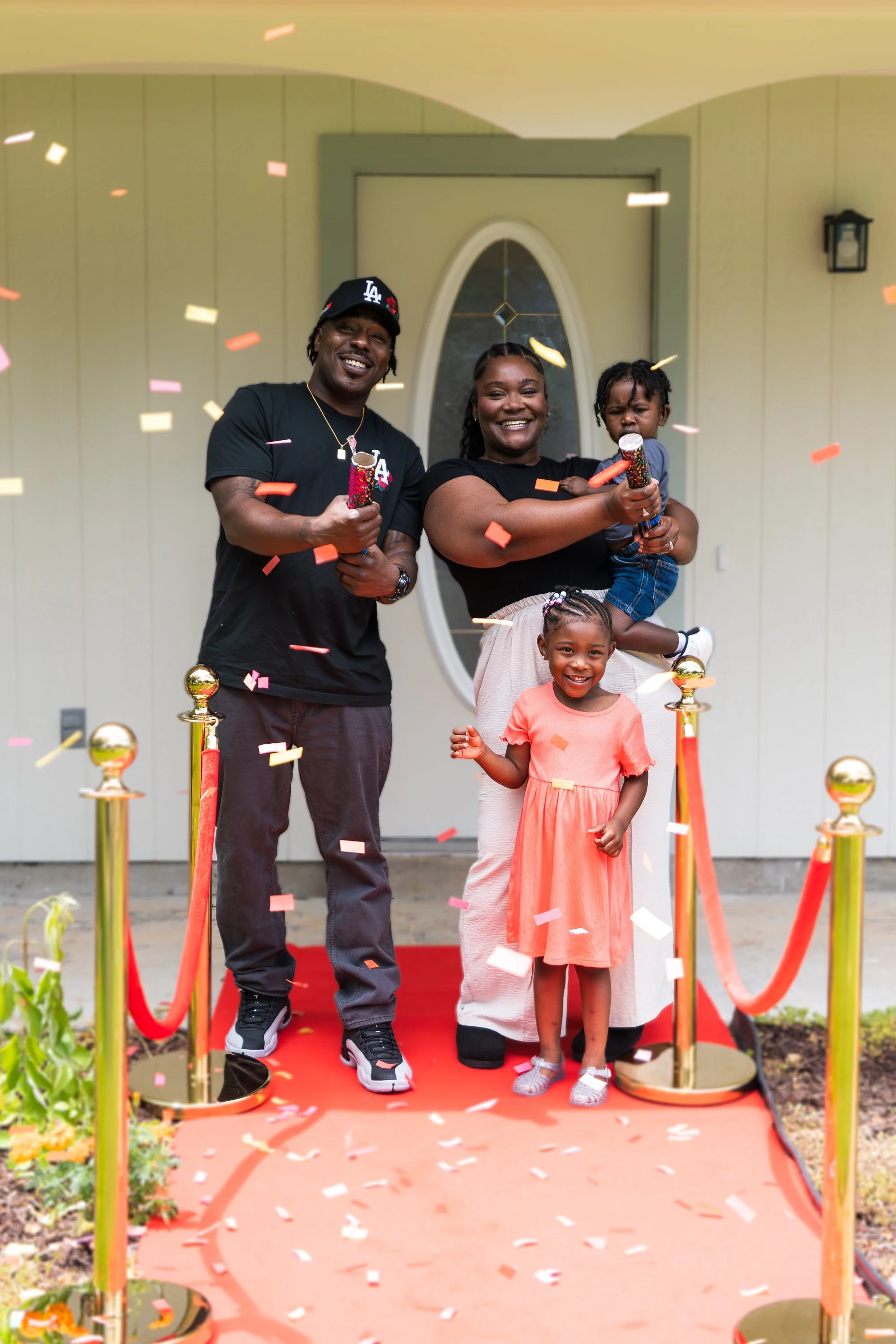A family celebrating a special occasion outdoors with confetti falling, standing on a red carpet at a doorway entrance, with two adults and two children smiling and holding party poppers.