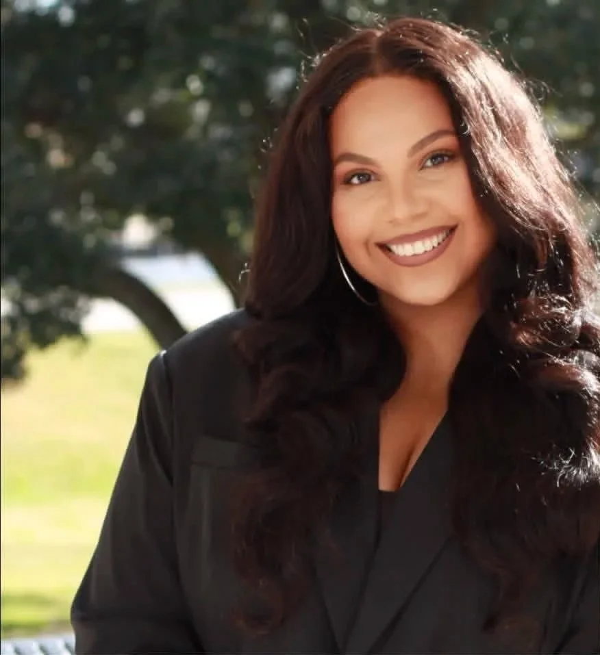 A woman with long curly dark hair smiling outdoors, wearing a black blazer and hoop earrings, with trees and sunlight in the background.