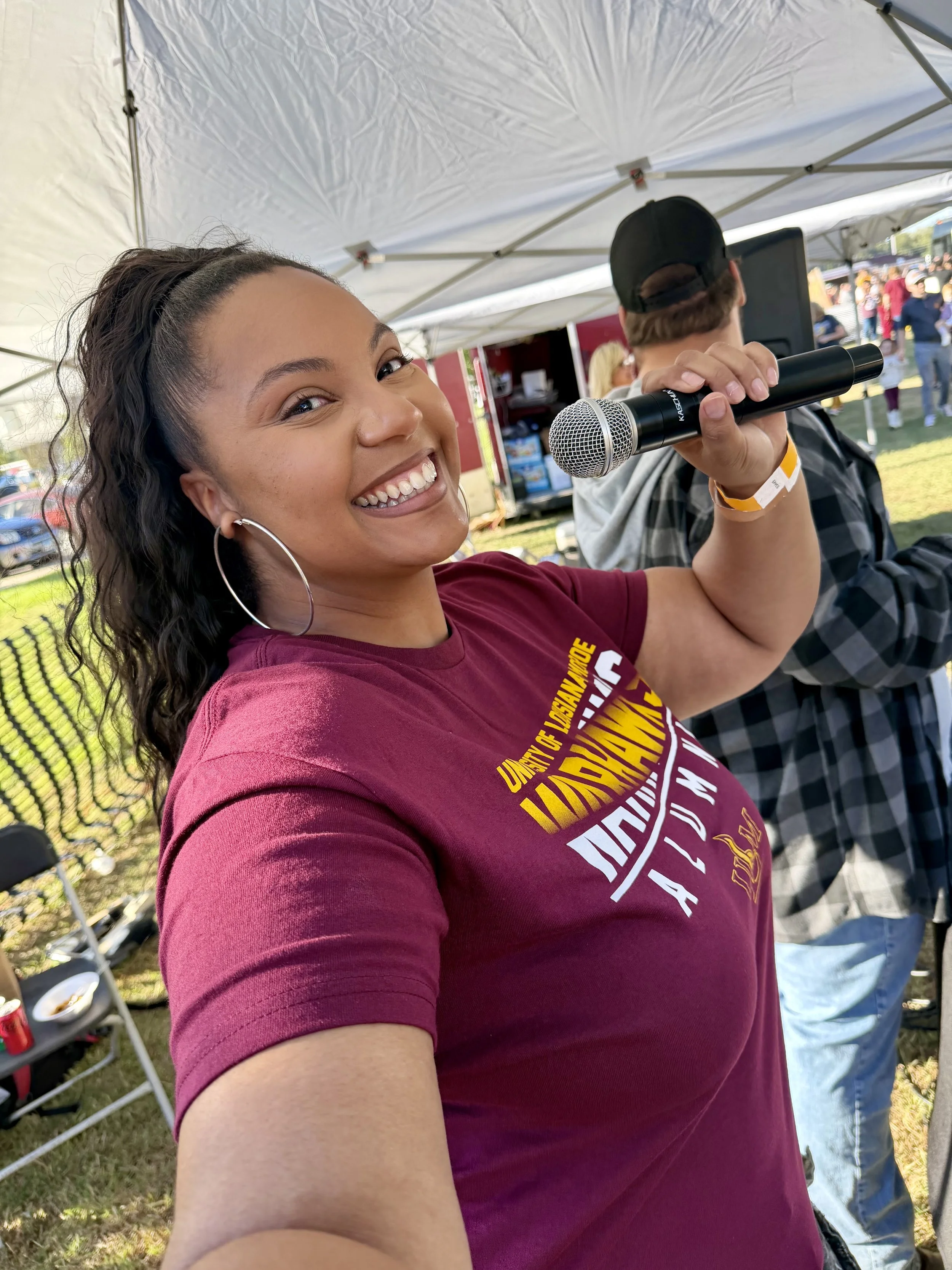 Young woman smiling and holding a microphone at an outdoor event under a white tent, with other people visible in the background.