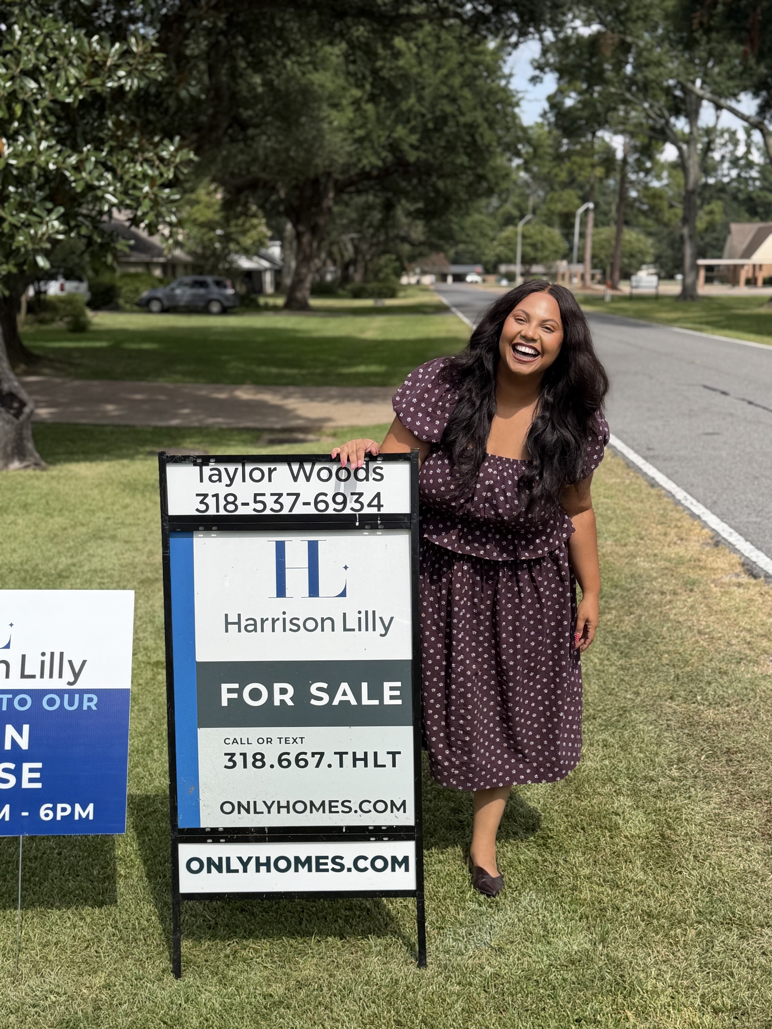 A smiling woman in a patterned dress standing outdoors on a grassy area holding a sign that says Harrison Lilly for sale with contact information, next to another sign related to homes, in a neighborhood with trees, houses, and a street in the background.