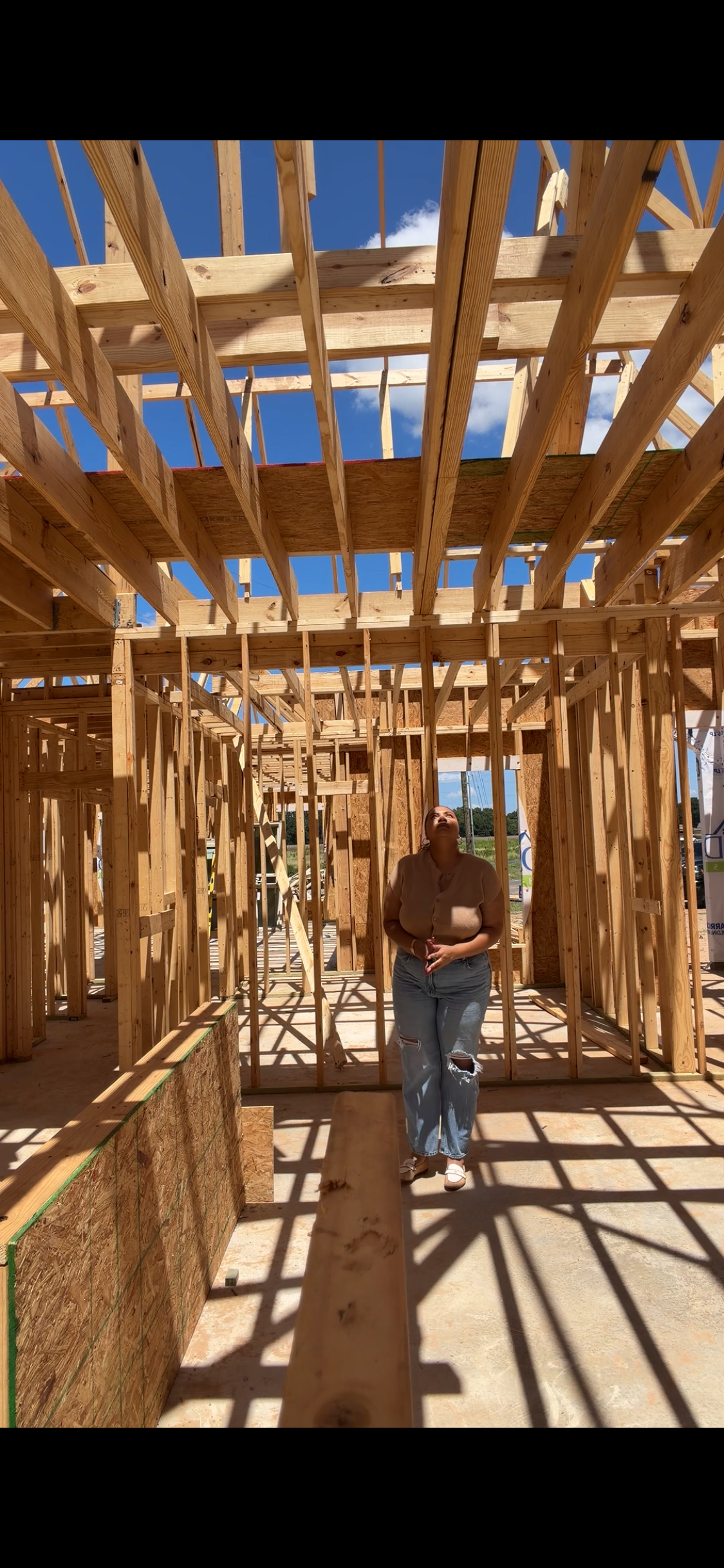 A woman in ripped jeans and a tan shirt standing inside a wooden house under construction, looking up at the blue sky with a few clouds.