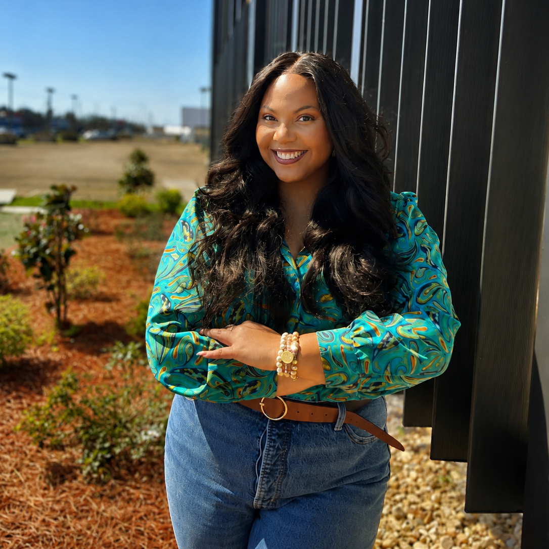A smiling woman with long dark hair, wearing a colorful patterned blouse and blue jeans, stands outdoors near a black wall, with a parking lot and clear blue sky in the background.