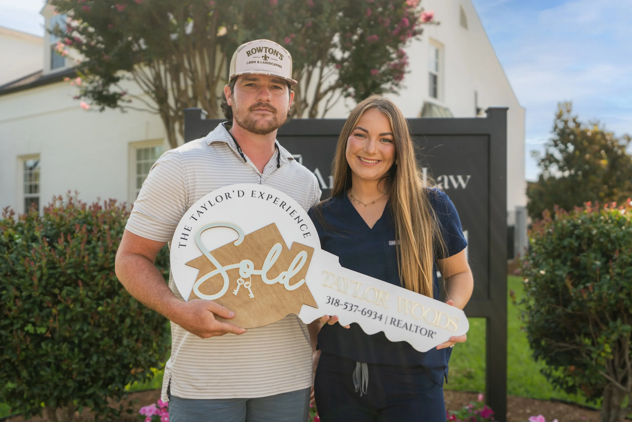 A man and woman standing outdoors in front of a tree and house, holding a large decorative key with the words 'The Taylor Experience' and contact information. The man is wearing a baseball cap and striped shirt, and the woman is smiling, wearing a navy blue scrub top.