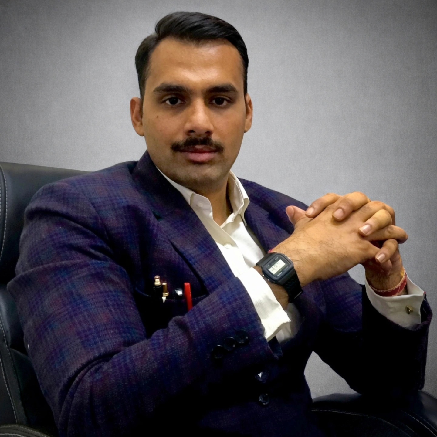 A man sitting in an office chair, wearing a dark suit and white shirt, with his hands clasped in front of him on the desk, looking at the camera.