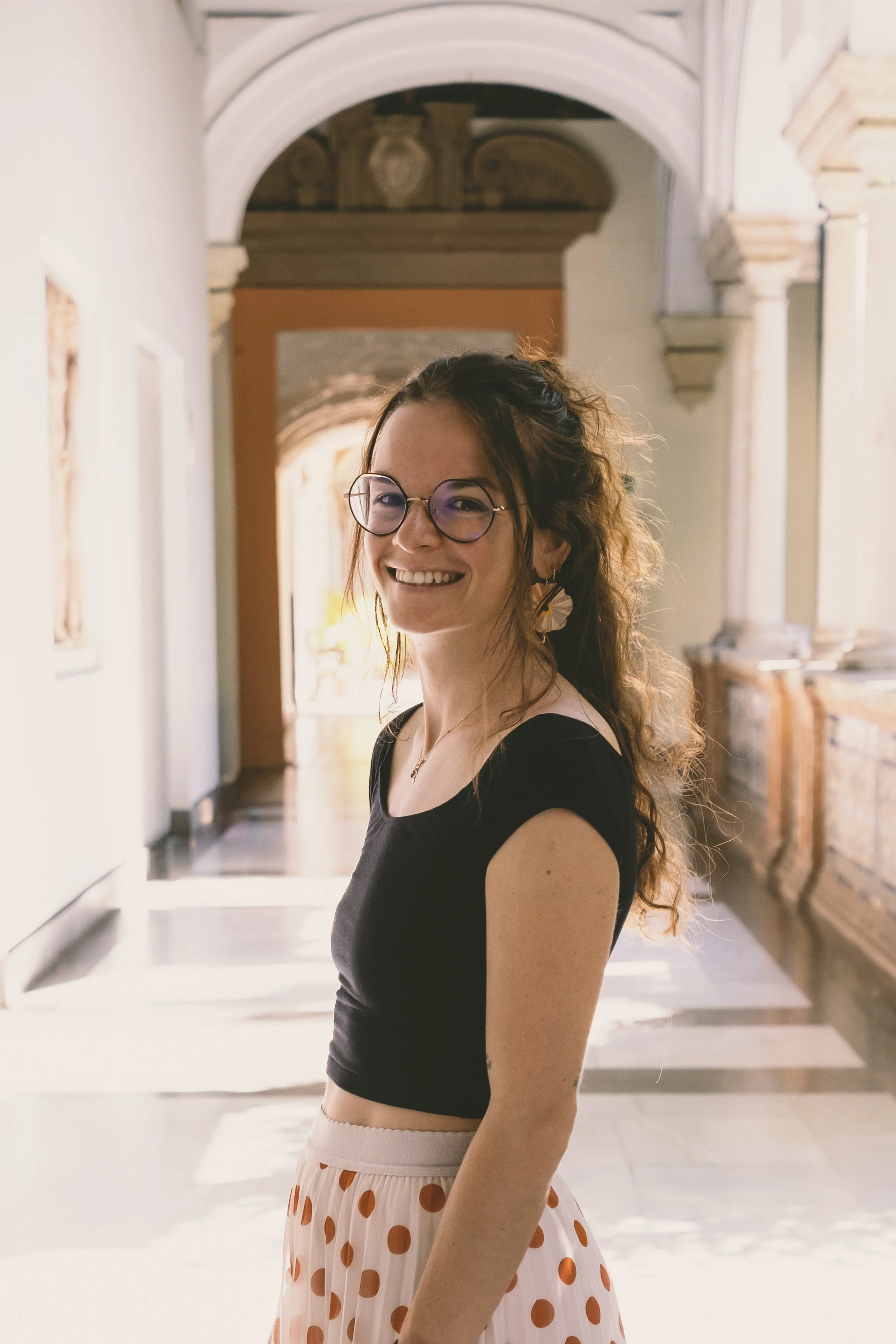 Jeune femme souriante avec lunettes, vêtue d'un haut noir et d'une jupe à pois orange dans un couloir avec architecture classique en pierre et plafond voûté.
