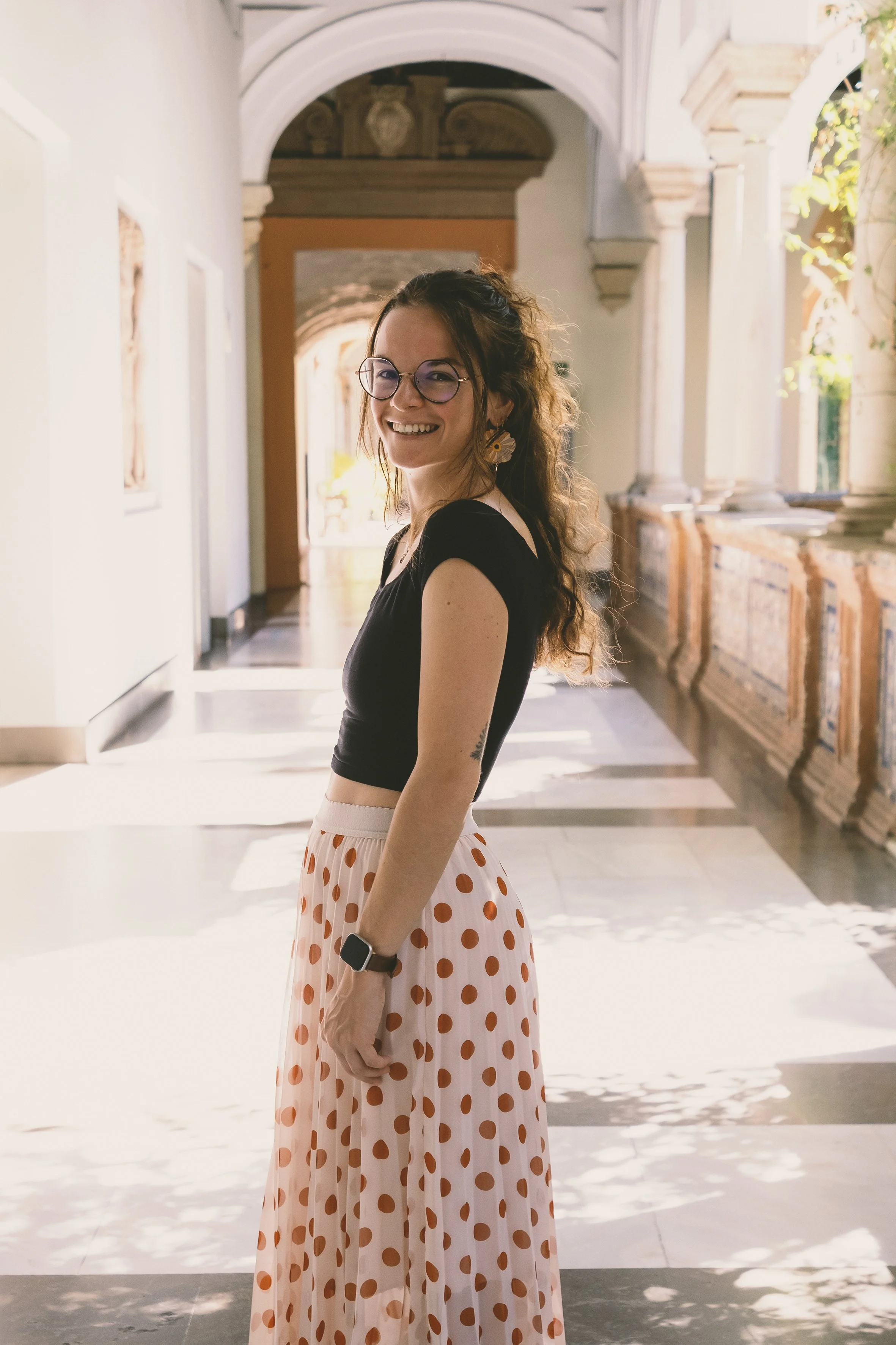 Femme souriante portant une robe à pois et des lunettes, dans un couloir lumineux avec des colonnes et des arches en architecture classique.