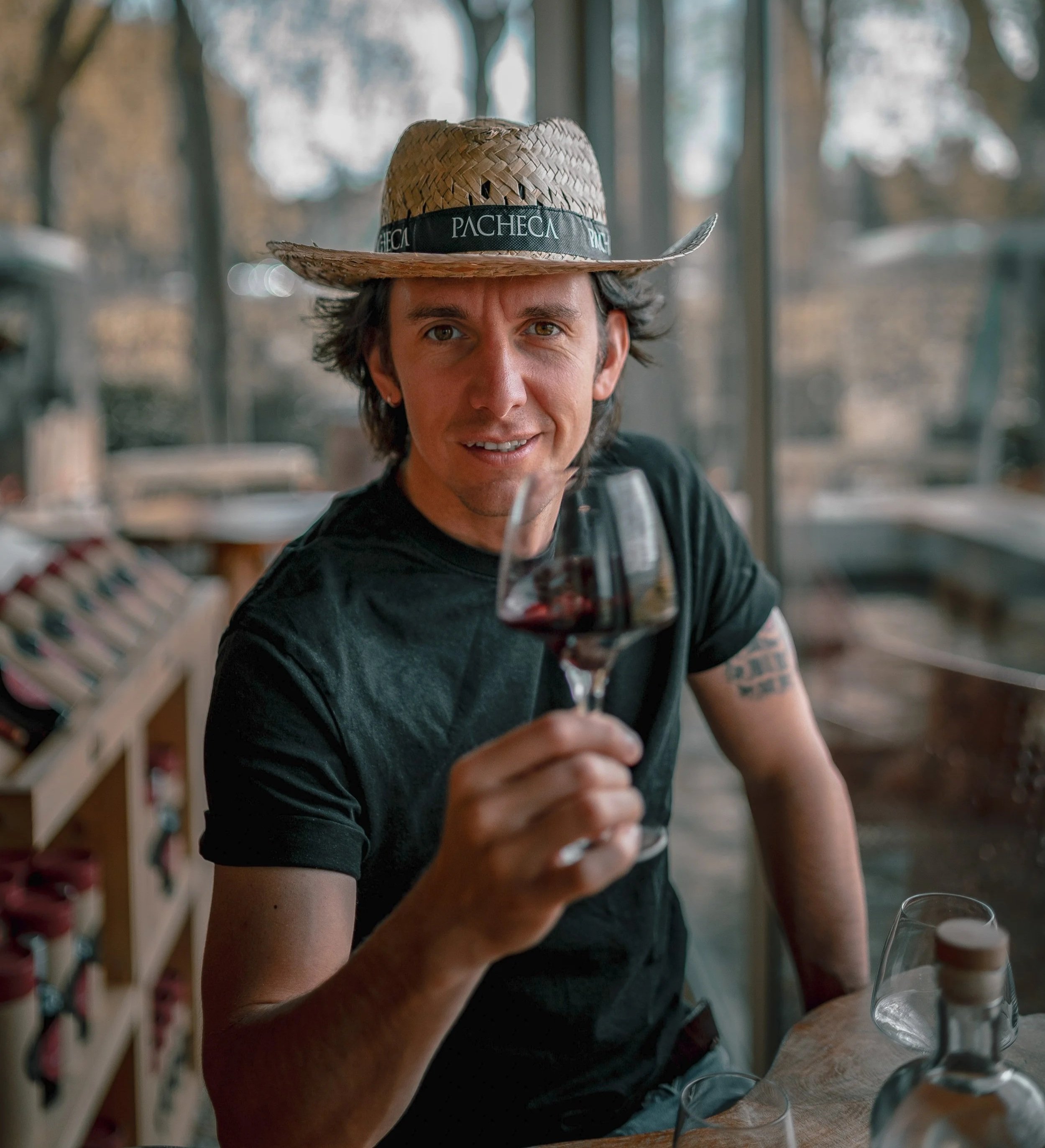 A woman with short dark hair wearing a straw hat with a black band that says 'PACHECA' is sitting in a restaurant or cafe, raising a glass of red wine towards the camera, with blurred wine glasses and shelves in the background.