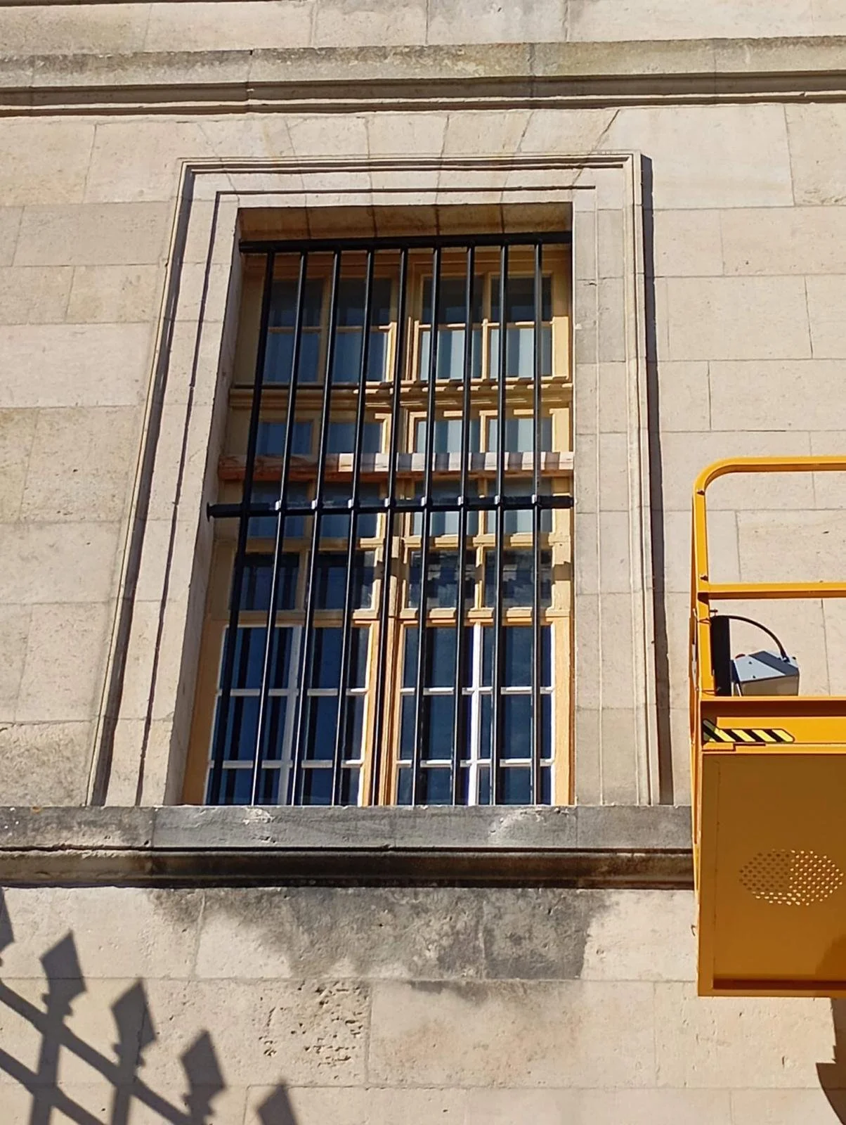 Façade en pierre avec une fenêtre à barreaux en métal et une grille en bois derrière. Partiellement visible, un escalier roulant jaune est à droite.