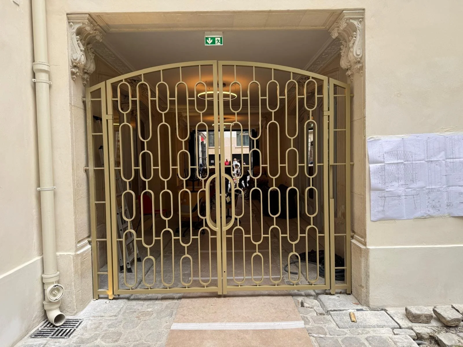 Une porte en métal ornée, située dans une entrée, avec un panneau de plan à droite et un panneau de sortie en haut. La porte donne sur un intérieur où des personnes et des vélos sont visibles.