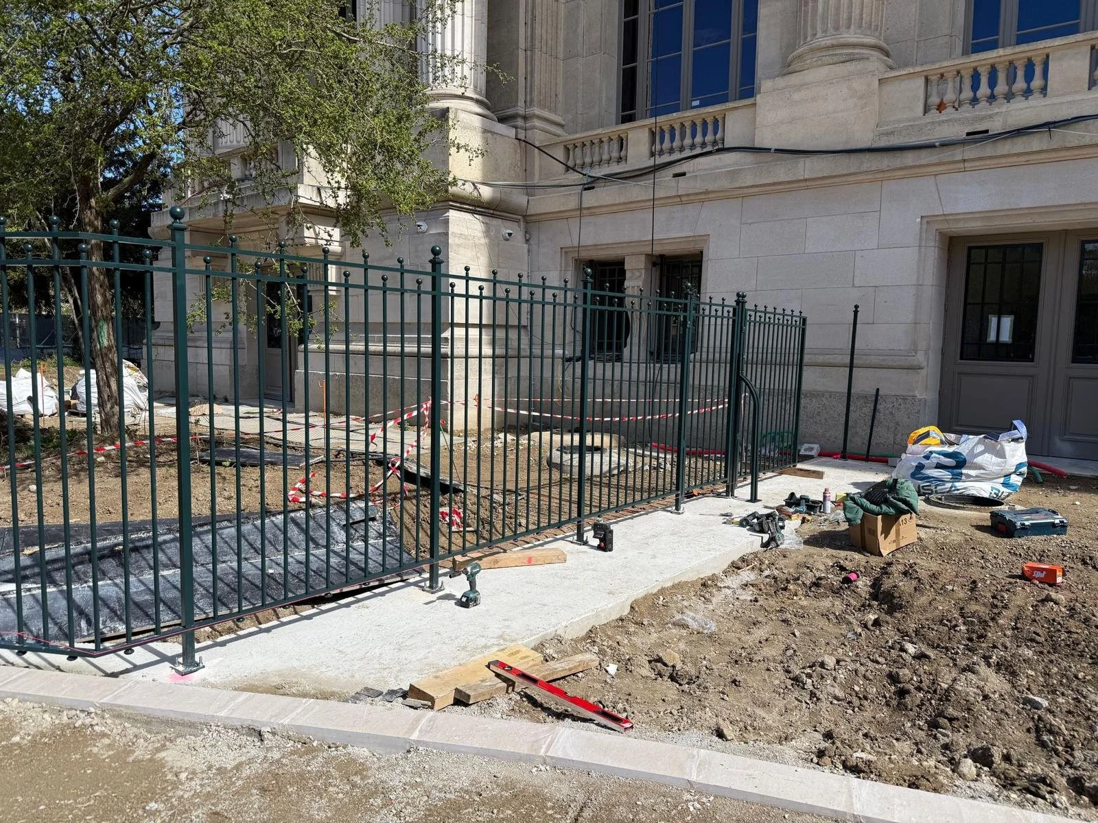 Travaux de clôture en cours devant un bâtiment en pierre, avec des outils et matériaux dispersés, et une partie du trottoir en construction.