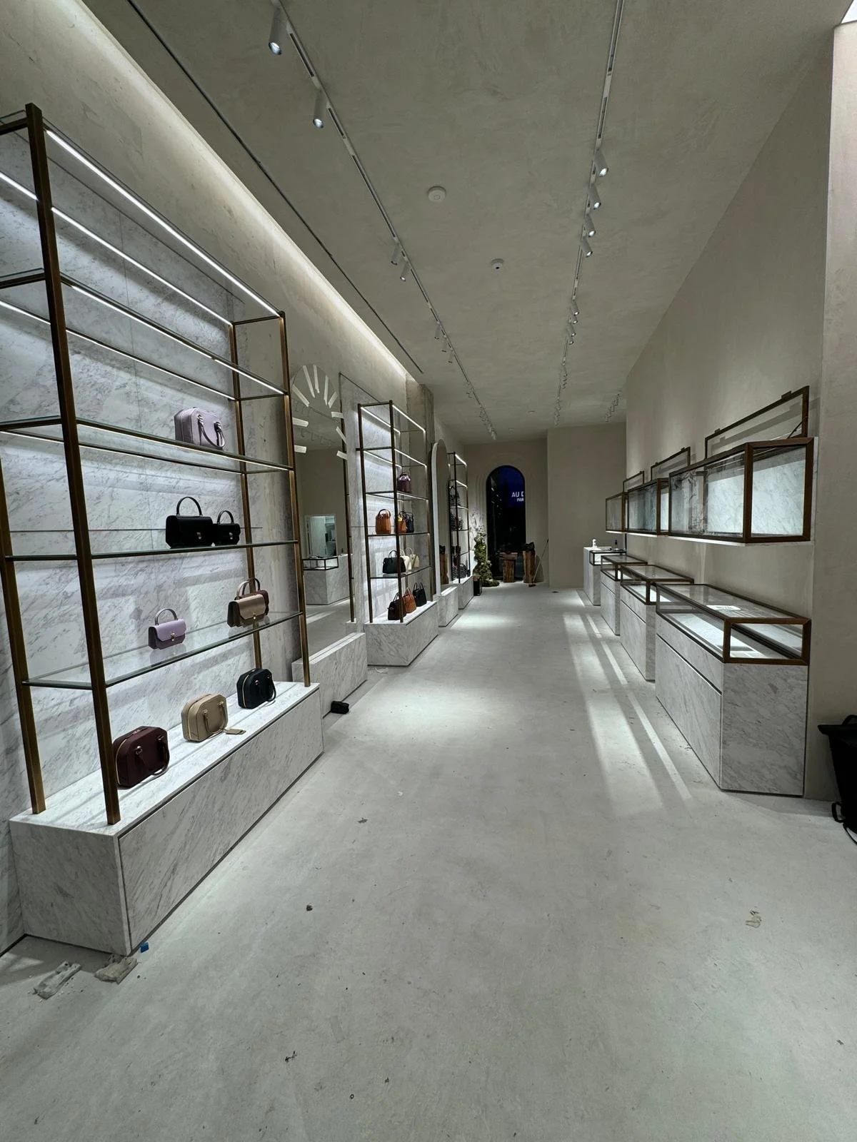 Une boutique élégante avec des étagères en marbre blanc et des cadres en métal doré, exposant des sacs à main de différentes couleurs, avec un sol en béton lisse et un plafond en béton avec un éclairage encastré.