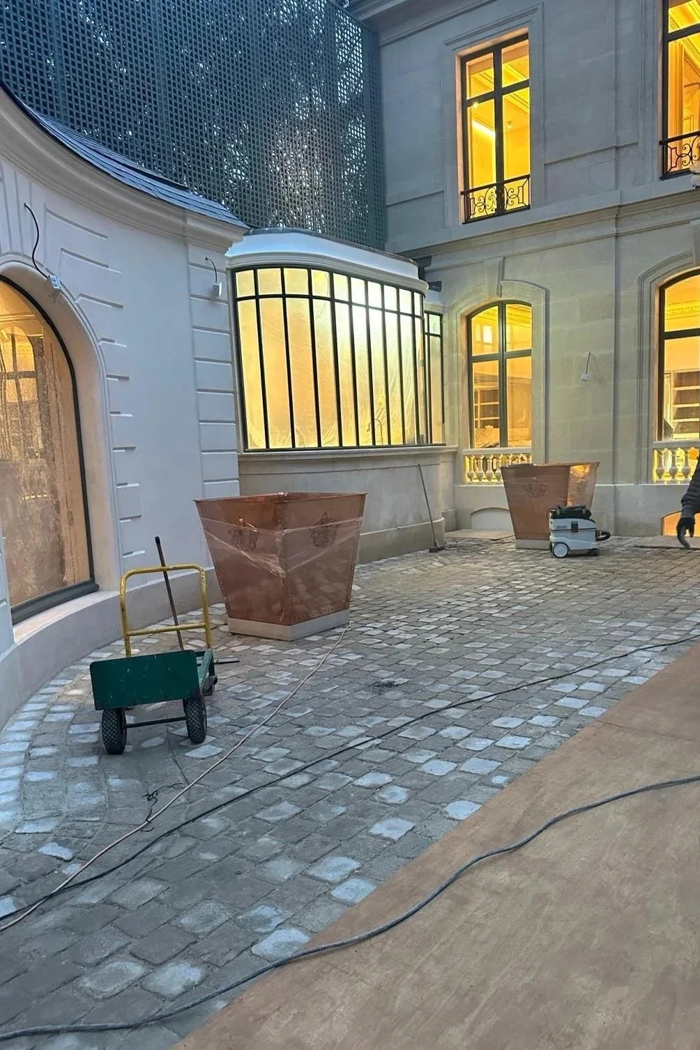 Cour intérieure d'un bâtiment en construction ou rénovation, avec des fenêtres éclairées en jaune, des plans de travail en cuivre, des outils et du câble électrique sur le sol pavé en pierre.