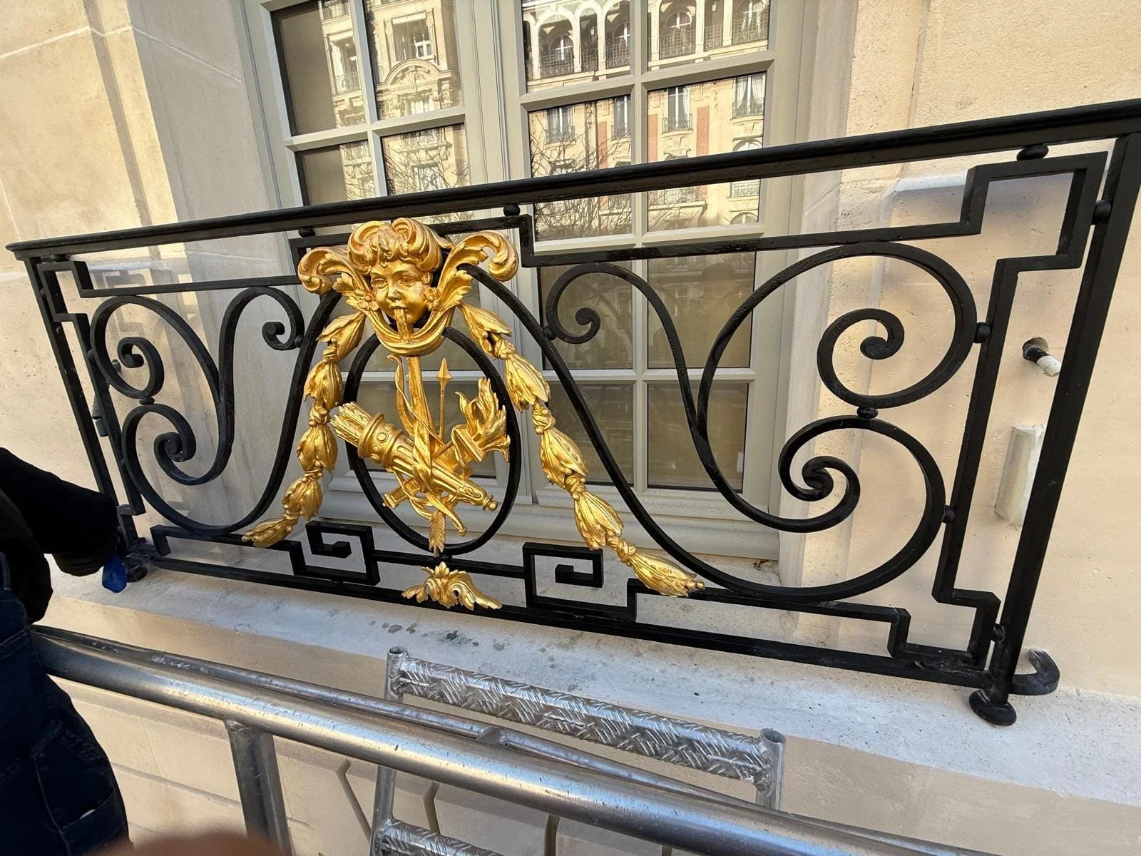 Délicate balustrade en fer noir et or avec une tête de ménade grimaçante, située devant une fenêtre en bois blanc à plusieurs carreaux, donnant sur une façade historique en pierre.