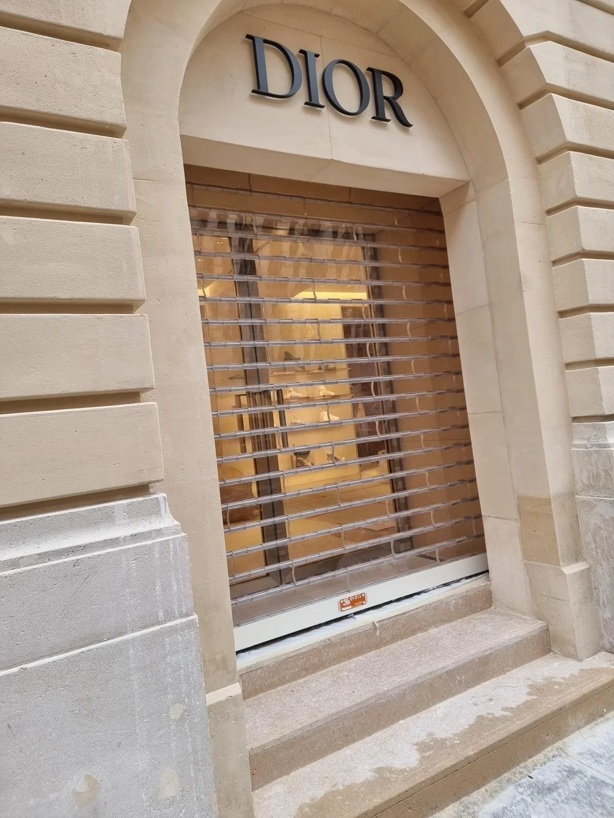 Vitrine d'une boutique Dior avec un store métallique fermant la vitrine, située dans un bâtiment en pierre avec pierre de taille, sur un trottoir en pierre.