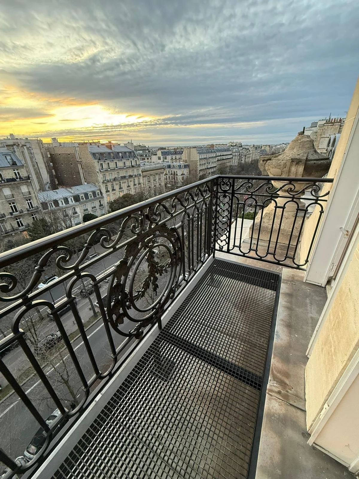 Balcon avec une grille en dessous et une rambarde en fer forgé, vue sur des immeubles parisiens et un ciel nuageux.