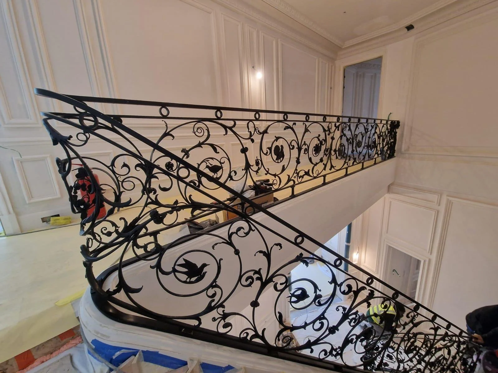 Balustre en fer forgé noir ornée de motifs floraux et de silhouettes de oiseaux, installé le long d'un escalier intérieur dans un espace décoré de murs en panneaux moulurés de couleur claire.