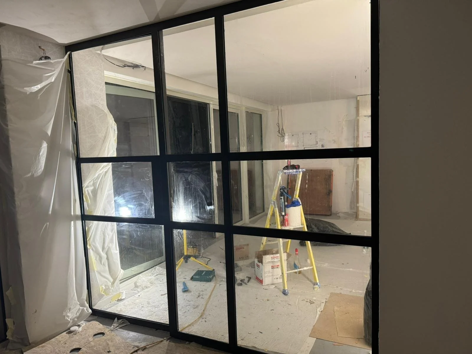 Une pièce en cours de rénovation vu à travers une grande fenêtre en verre divisée en plusieurs sections par une grille métallique.