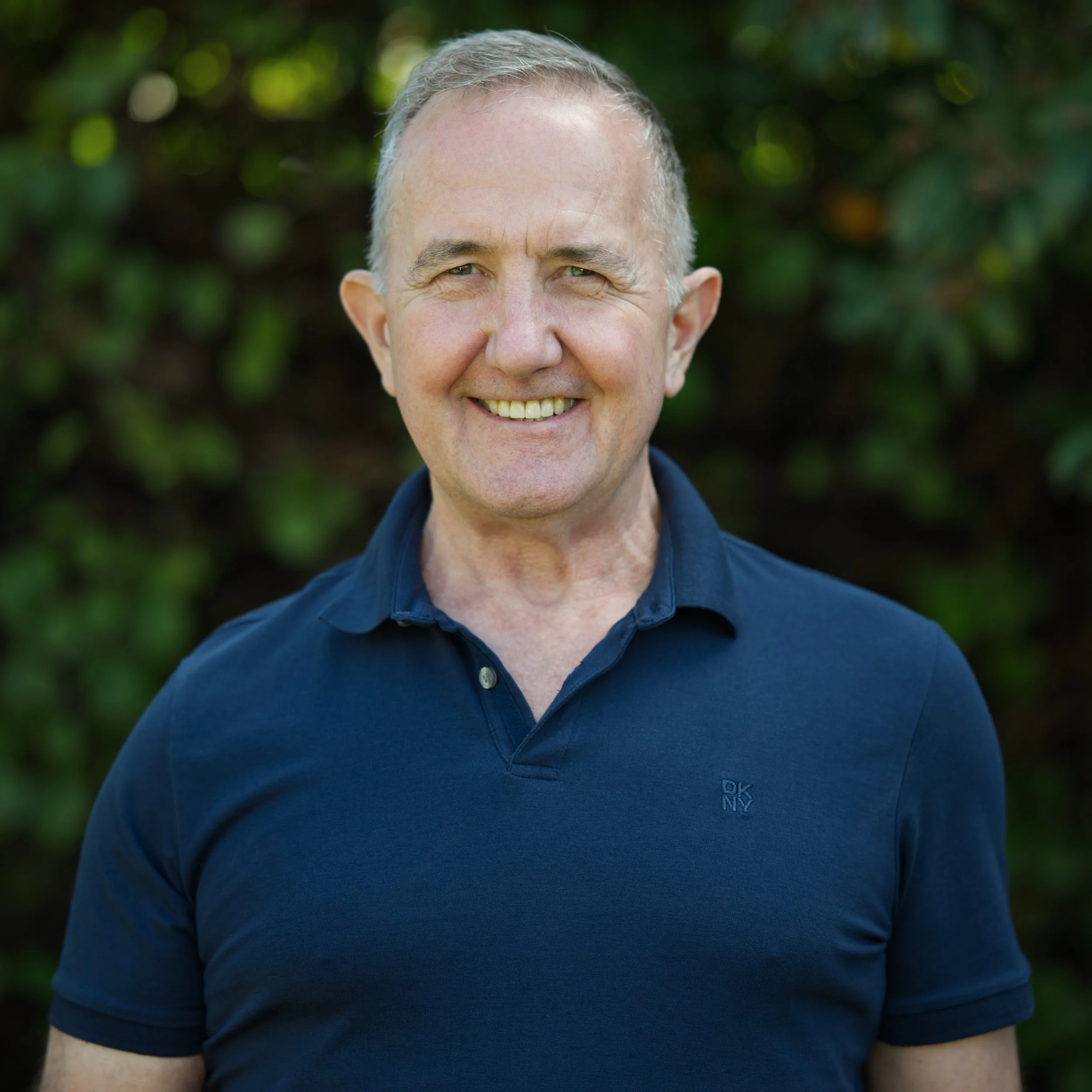 Portrait of photographer Chris Angel smiling outdoors, wearing a navy blue polo shirt against a softly blurred green background.