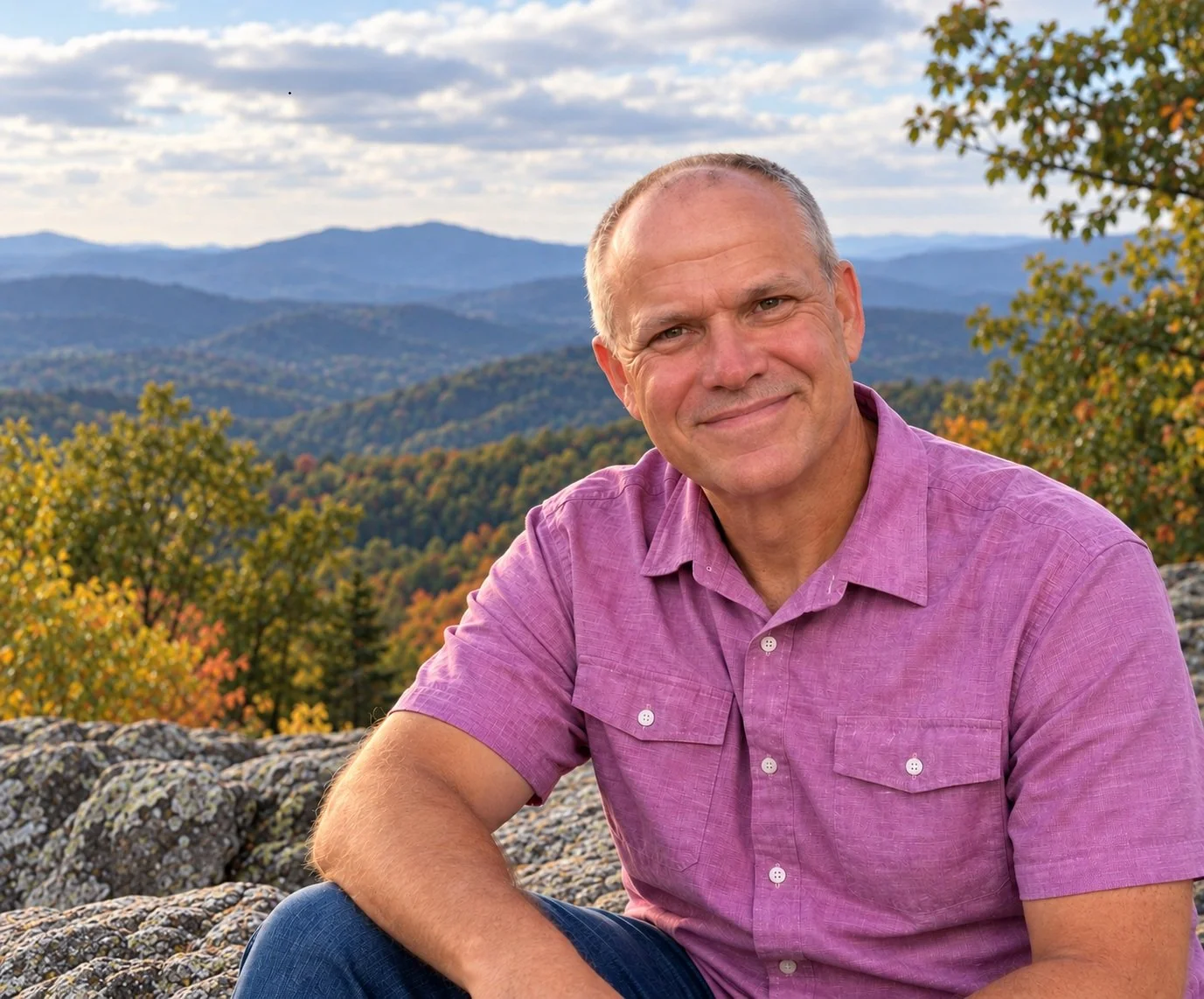 A smiling man in a pink shirt sitting outdoors on rocks with a mountain landscape and autumn trees in the background.
