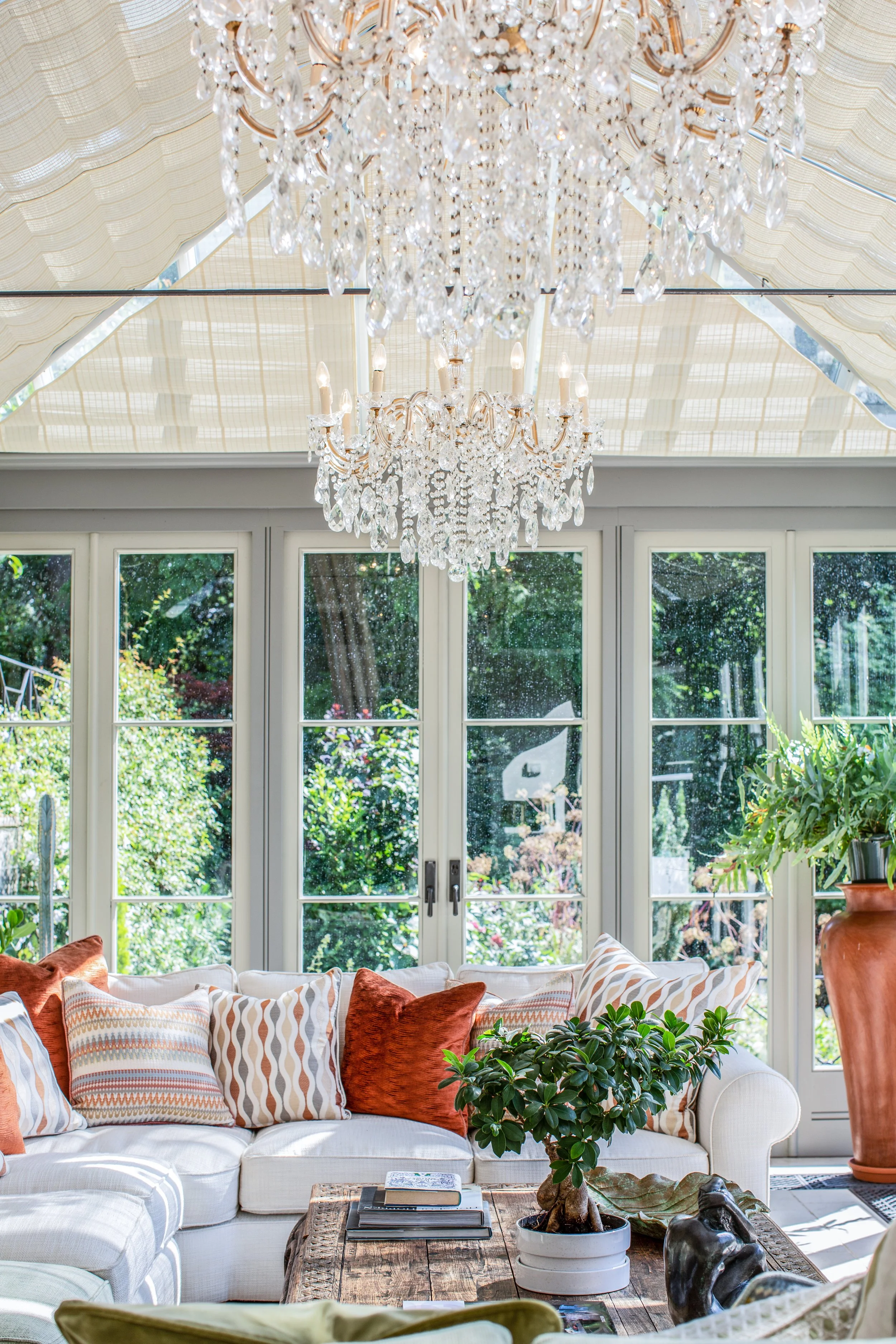 A cozy living room with a white sectional sofa decorated with patterned and solid-colored pillows, a rustic wooden coffee table with books and decorative objects, large windows with a view of green trees, and two elegant crystal chandeliers hanging f