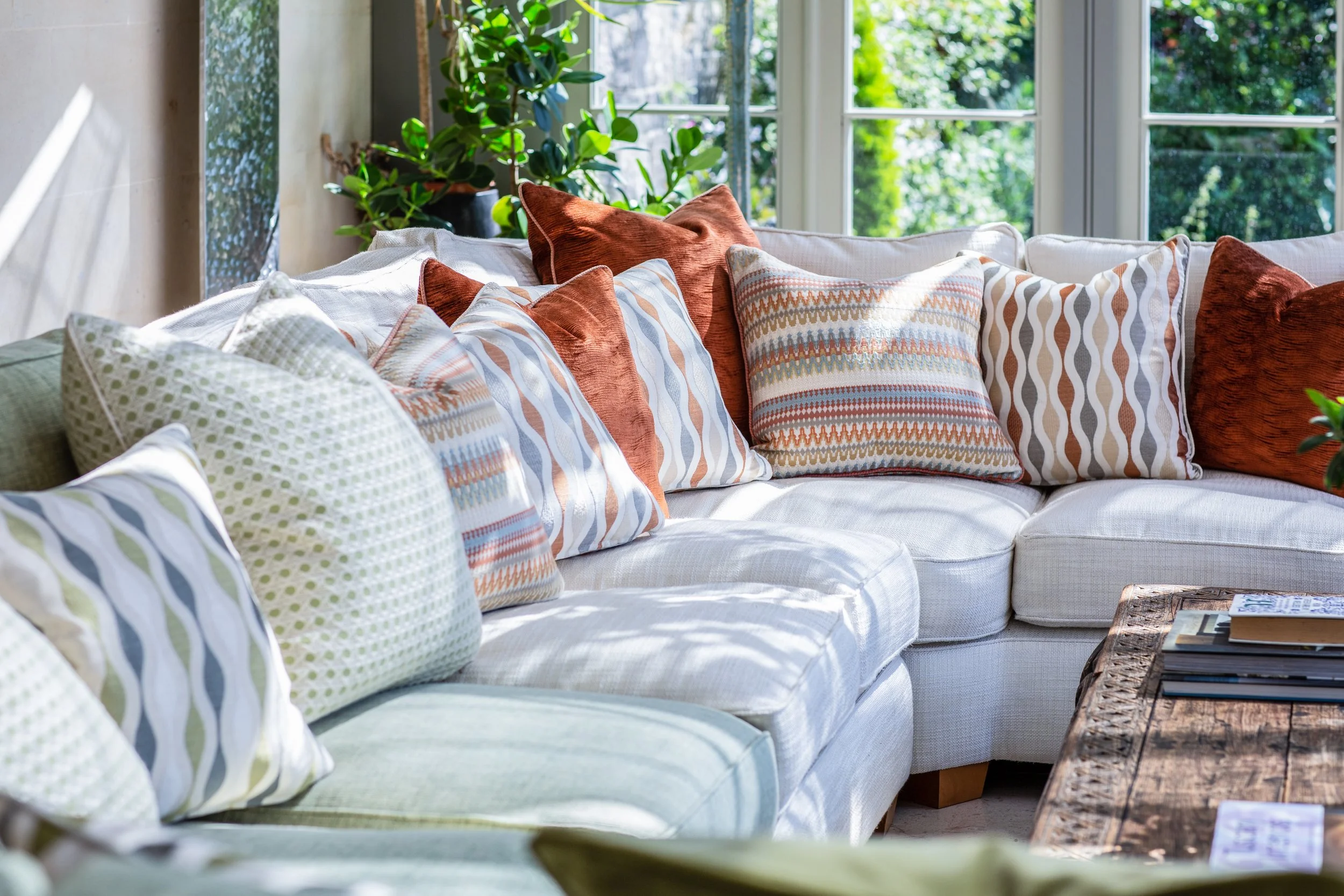 A cozy living room area with a white sofa decorated with various patterned and rust-colored throw pillows, a wooden coffee table with magazines, and large windows allowing natural light to fill the space. Green houseplants sit near the window, adding