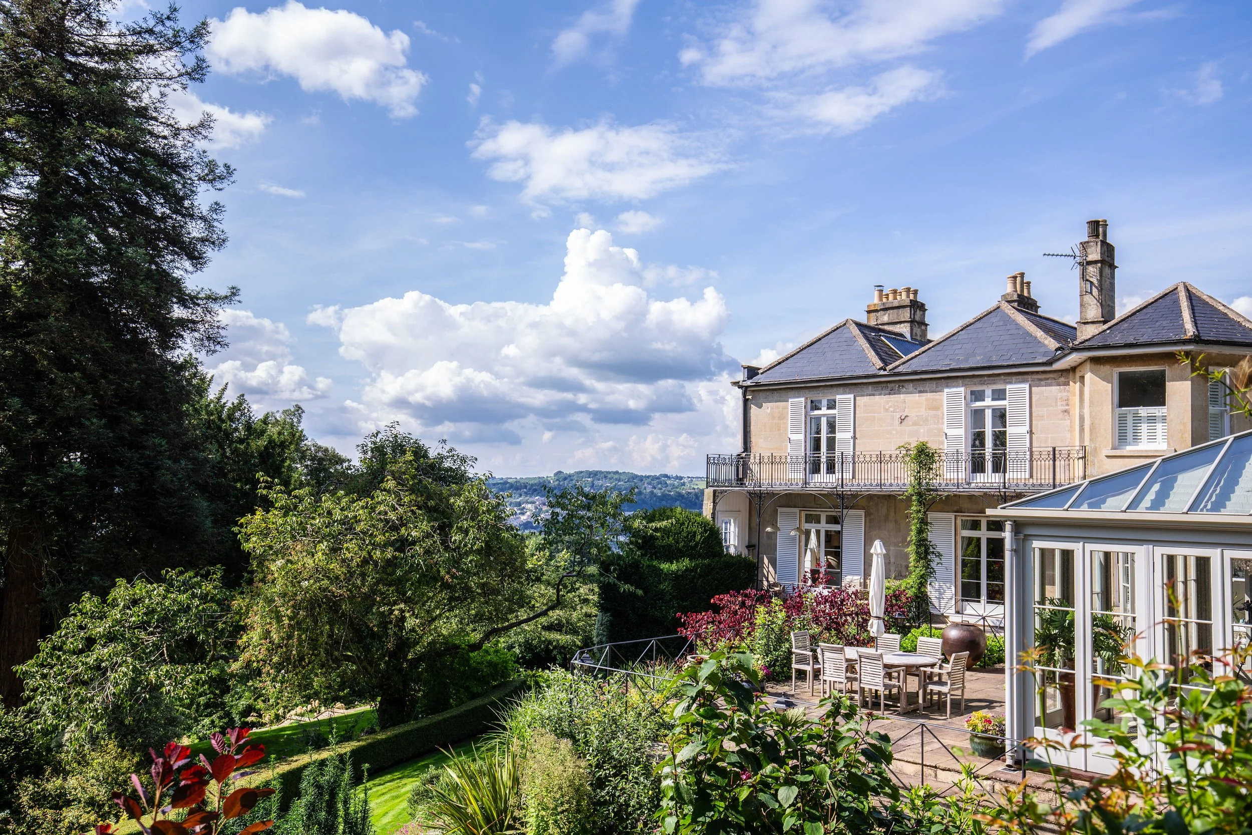 A large house with gray slate roof and white shutters on windows, surrounded by a lush garden with trees, plants, and flowers, under a partly cloudy blue sky.